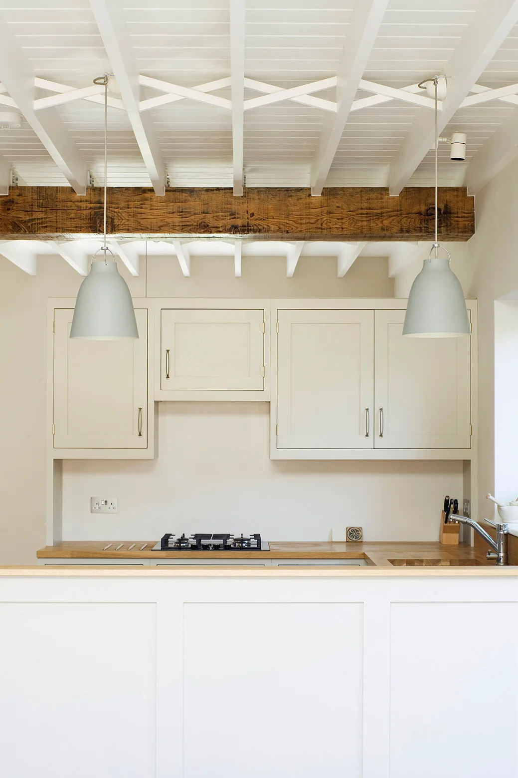 A bright kitchen features cream cabinets, a wooden countertop, two gray pendant lights, and an exposed wooden beam across a white ceiling.