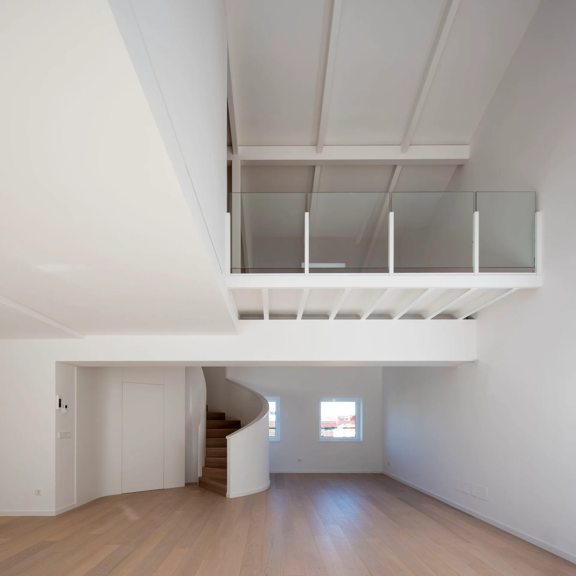 An empty modern room with light wood floors, a curved staircase, and a white mezzanine level featuring glass railings.