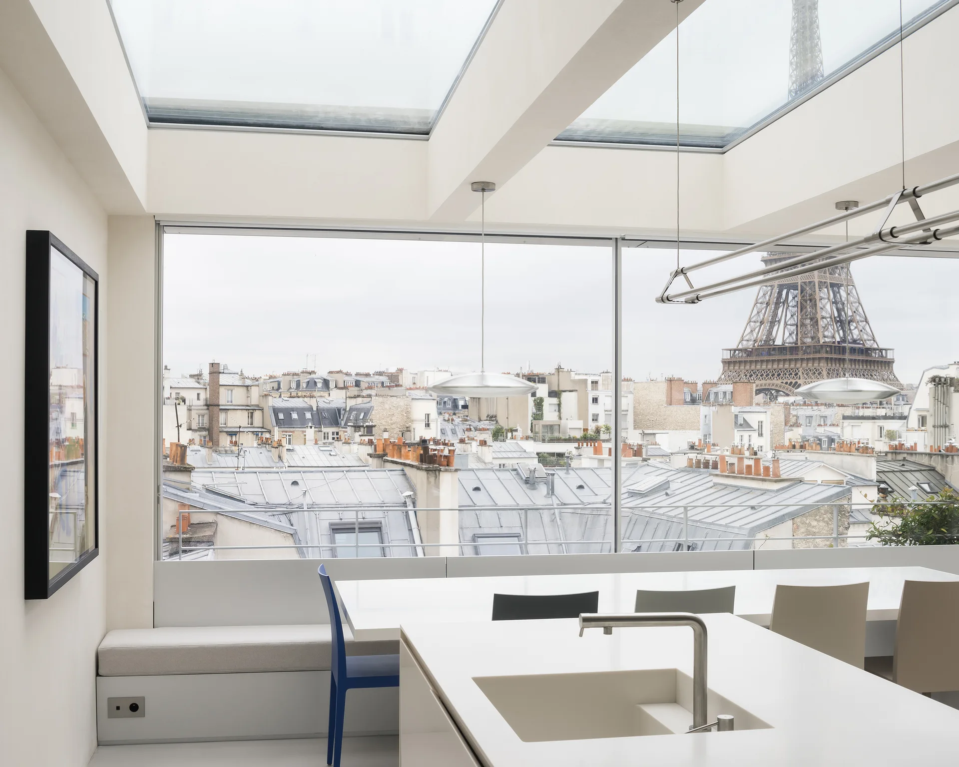 A minimalist white kitchen featuring an integrated island sink, a built-in bench, and a large horizontal window looking out over the Paris cityscape.