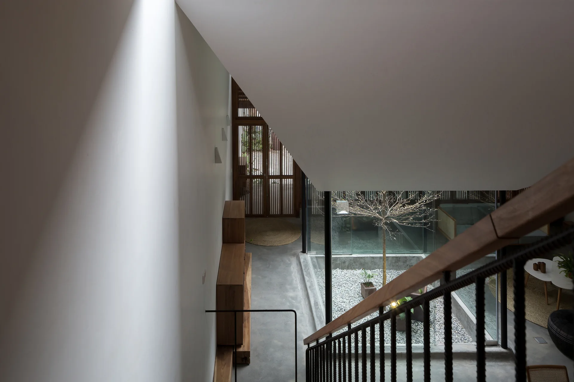 Looking down a modern staircase with a dark metal railing toward a grey concrete floor, a wooden cabinet, and a glass-enclosed interior gravel courtyard.