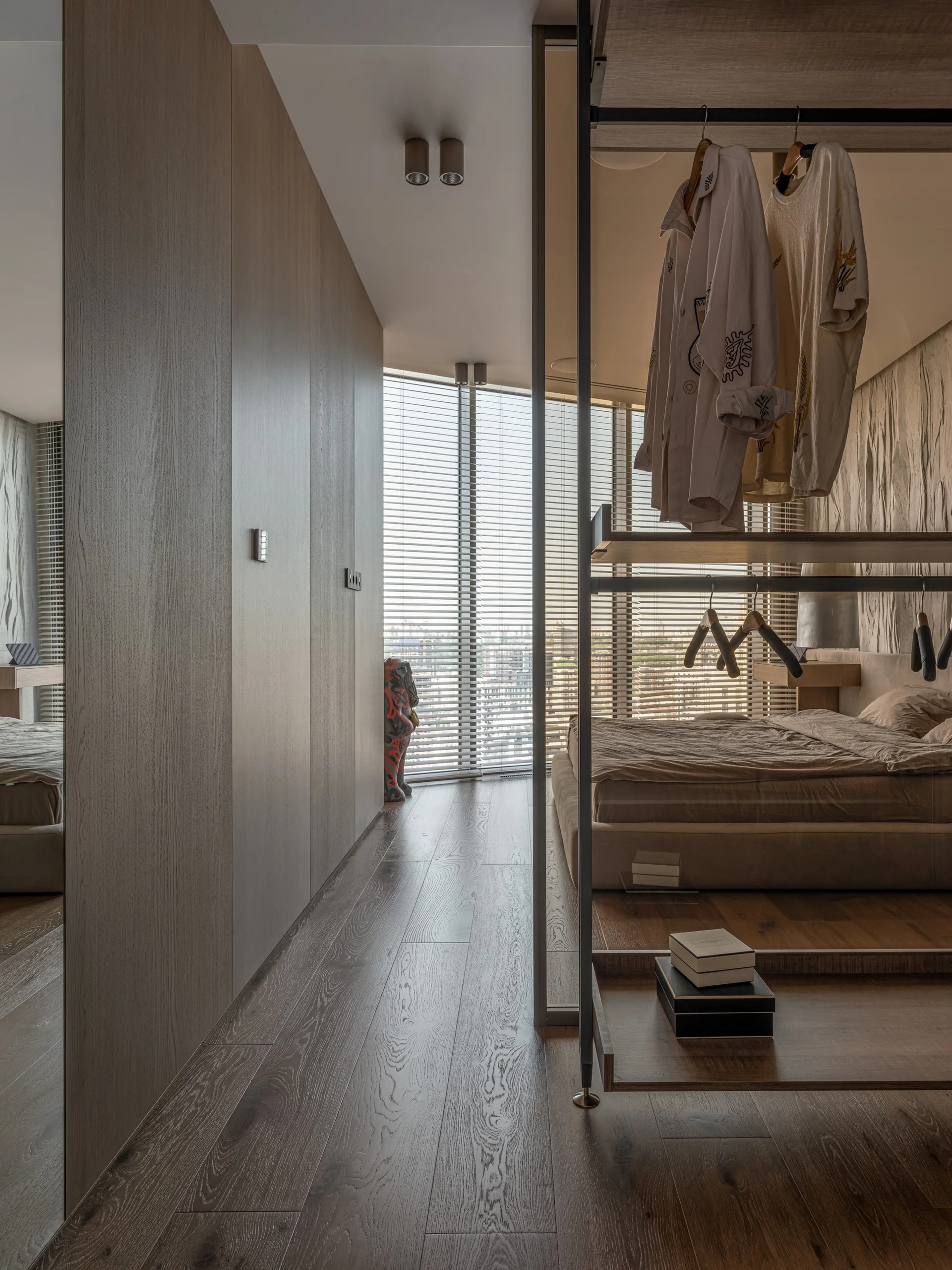 A modern minimalist bedroom featuring a low-profile bed, an open metal clothing rack, wood wardrobe panels, and window blinds.