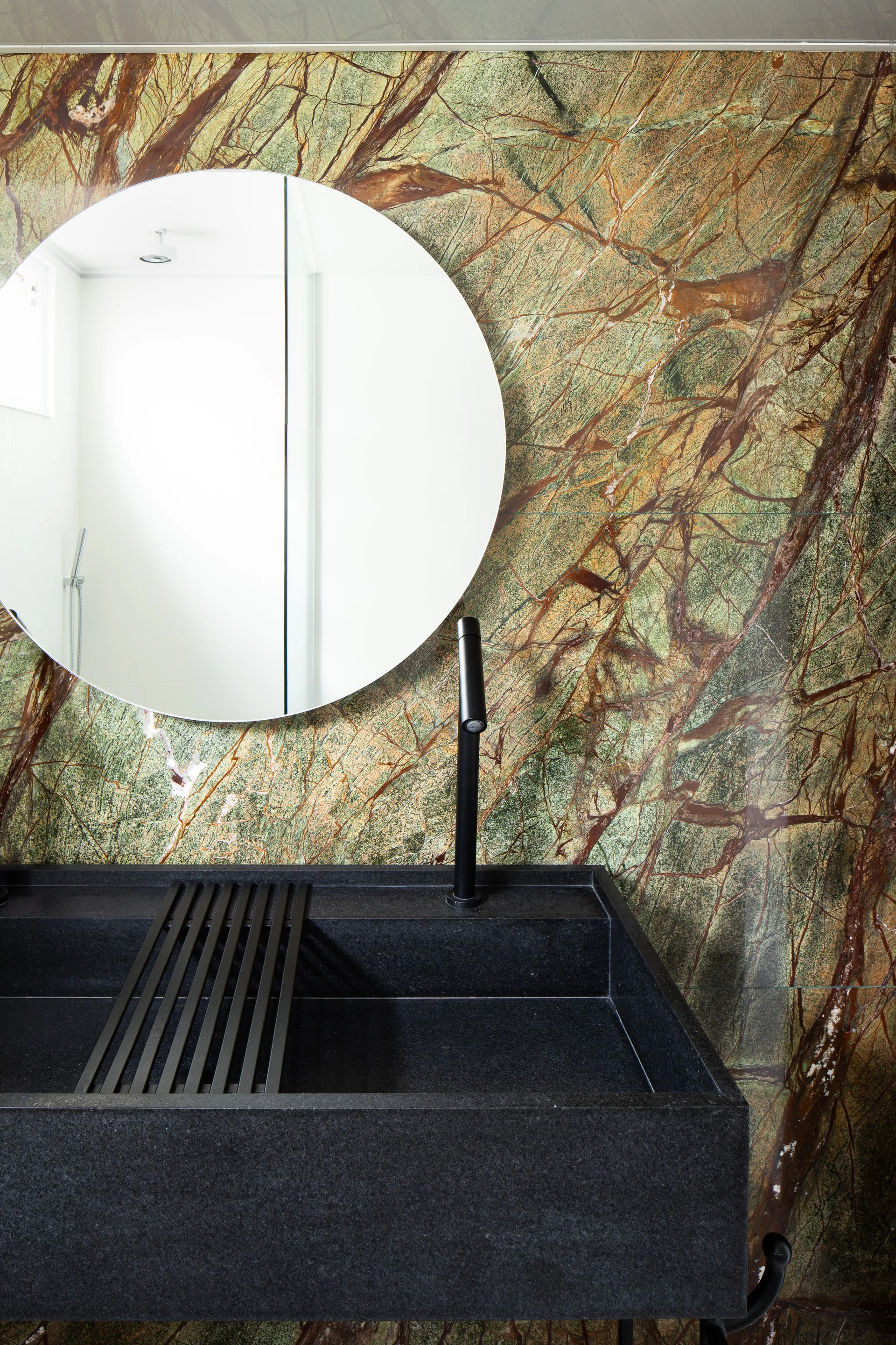 A bathroom features a black stone vanity sink with a tall faucet set against a green veined marble wall and a round mirror.