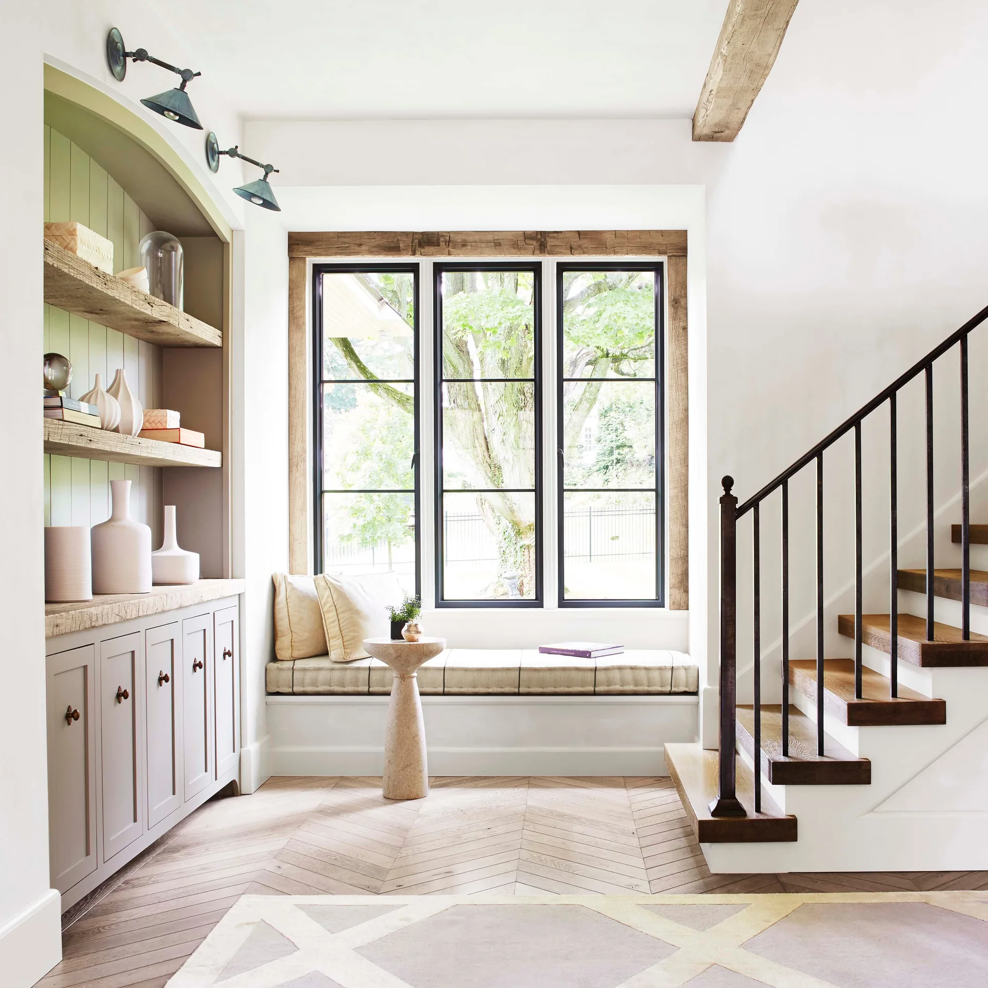 A bright room features a built-in window seat under a black-framed window, next to an arched shelving unit and a wooden staircase.