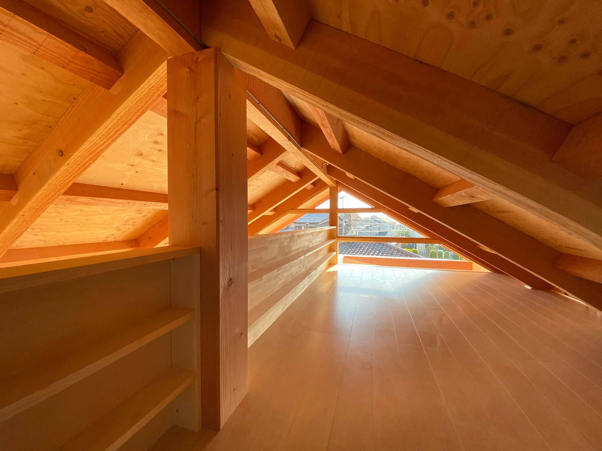 A bright attic interior constructed entirely of light wood, featuring exposed structural beams, plank flooring, and a small triangular window.