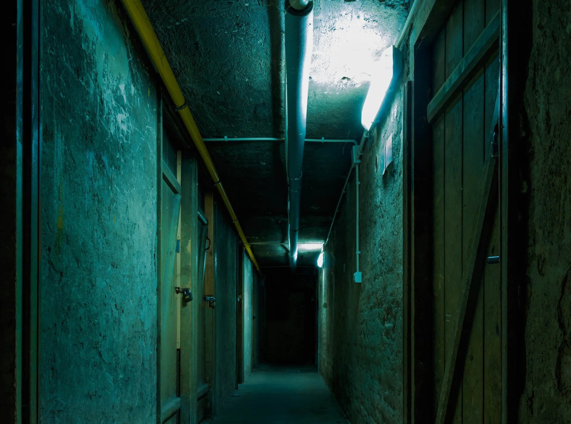 A dimly lit, narrow industrial basement hallway features textured plaster walls, several wooden doors with padlocks, and exposed ceiling pipes.