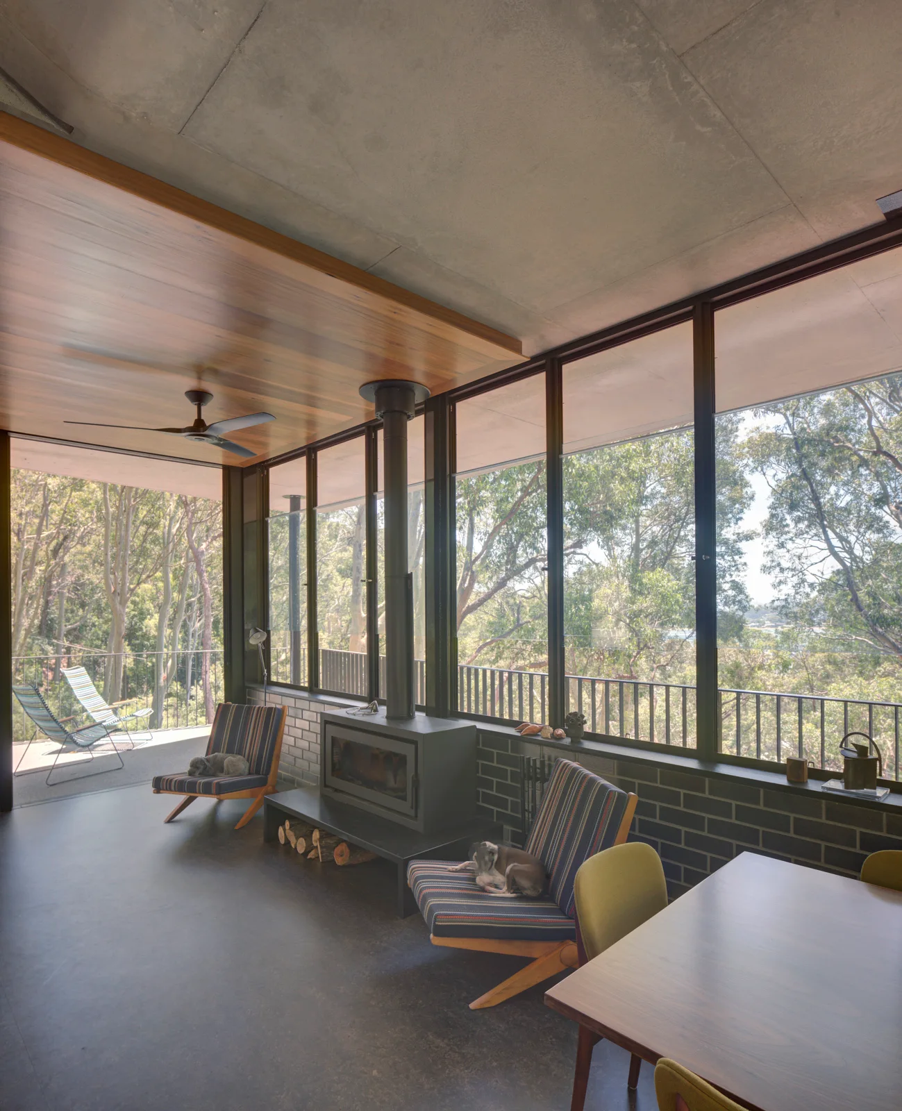 A modern living room features striped armchairs, a black wood-burning stove, and a wall of windows looking out onto a dense forest.
