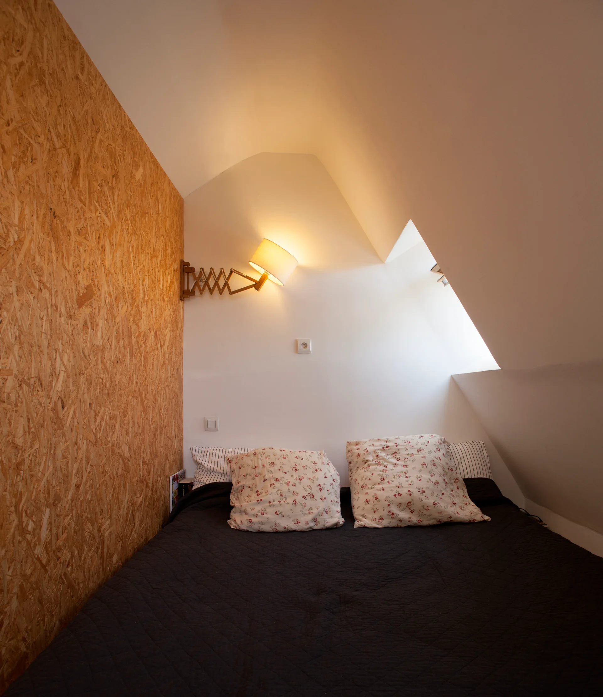 An attic bed nook features a dark-covered bed beneath a sloped ceiling next to an oriented strand board accent wall with a wooden scissor lamp.