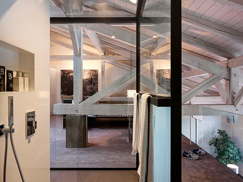 A modern attic bathroom features a tiled shower with a black-framed glass wall looking into a bedroom with exposed wooden ceiling trusses.