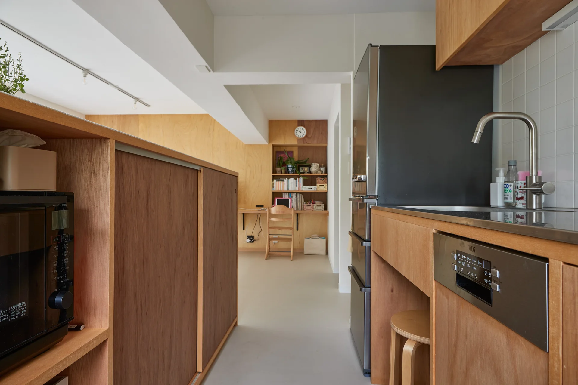 A galley kitchen with light wood cabinetry and a stainless steel sink looking toward a compact study nook with built-in wooden shelves.