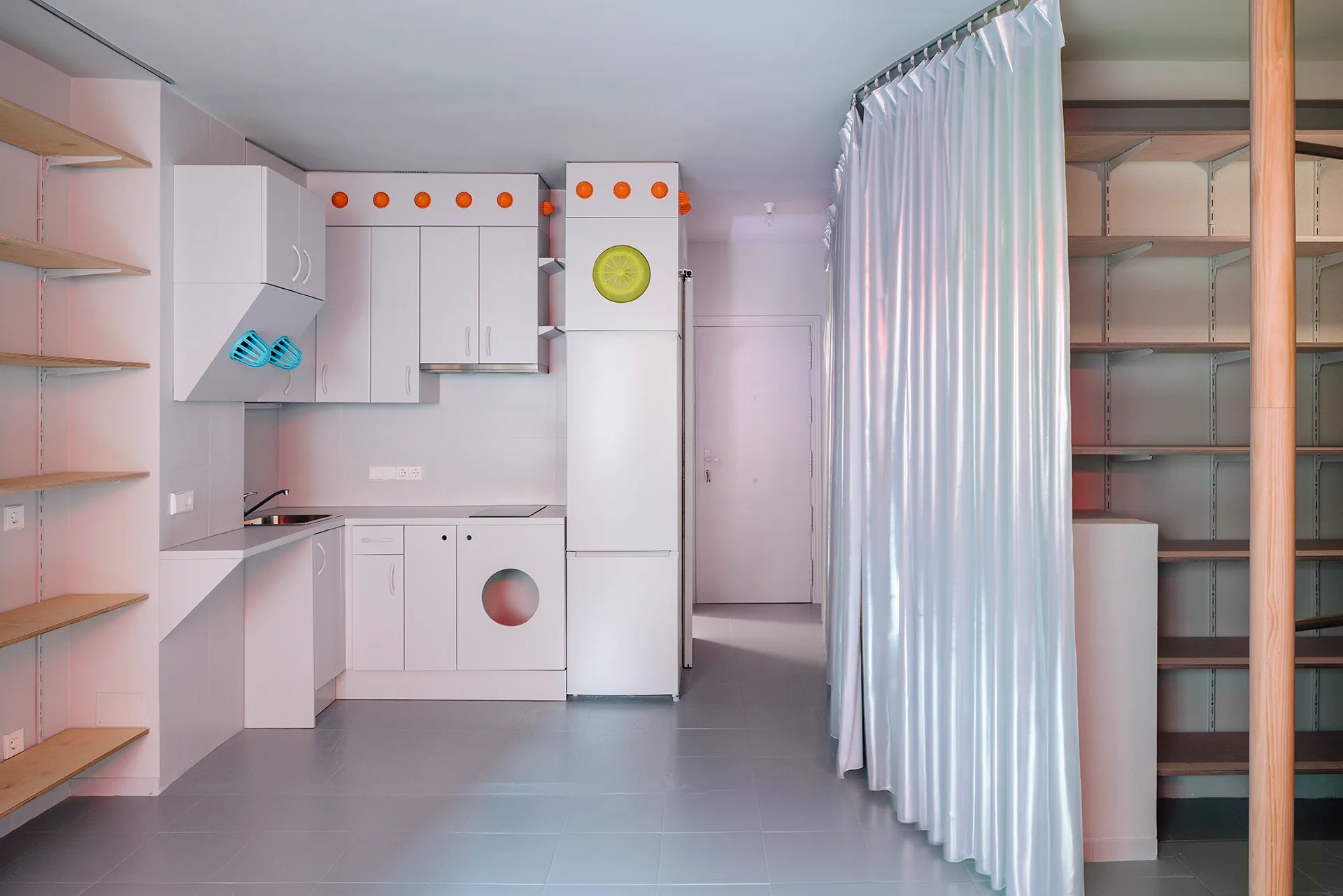 A minimalist white studio kitchen features modular cabinetry, an integrated washing machine, open wood shelving, and a tall shimmering silver curtain divider.