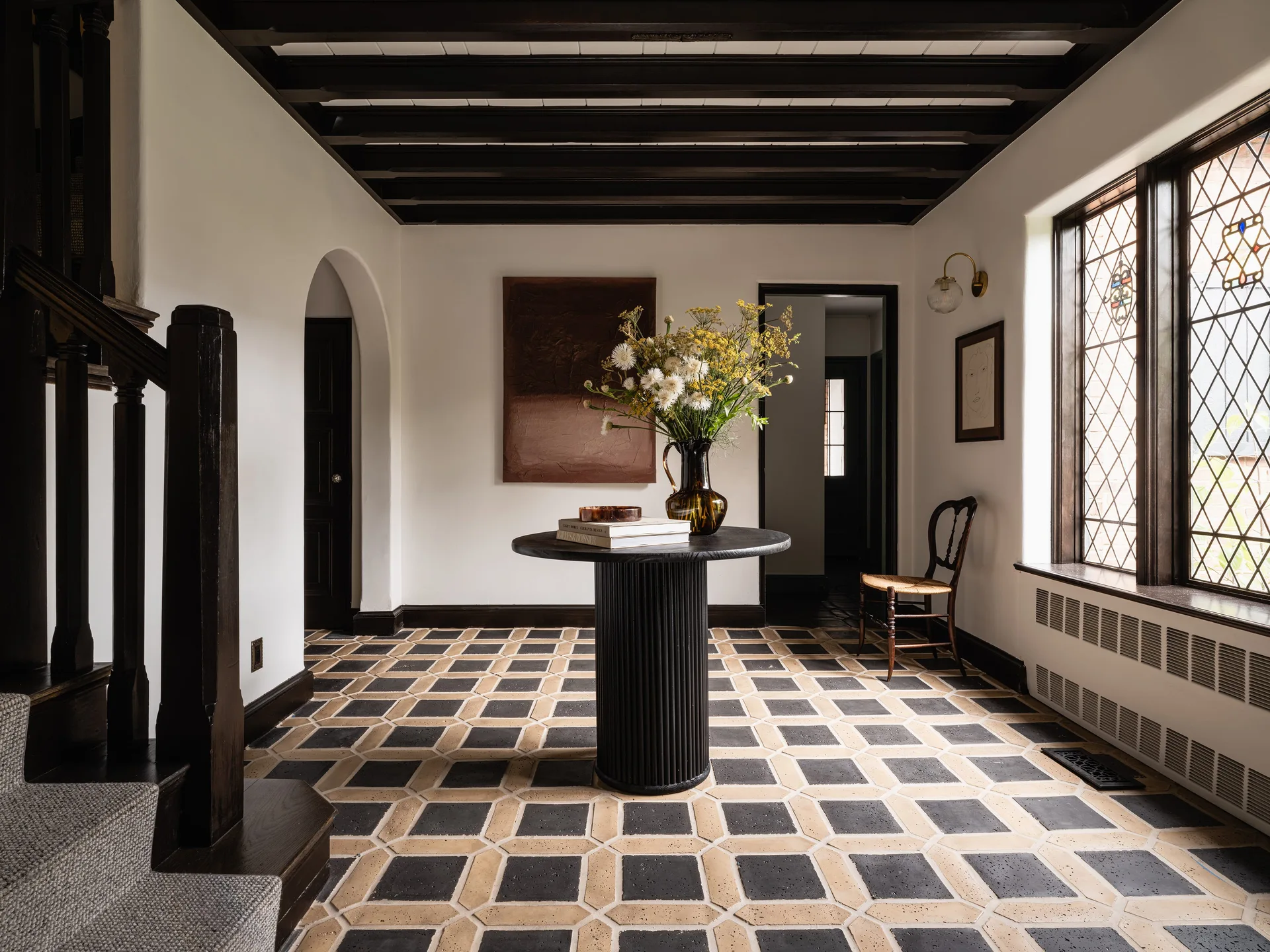 An entry foyer features a black fluted pedestal table, geometric floor tiles, and dark wooden ceiling beams.