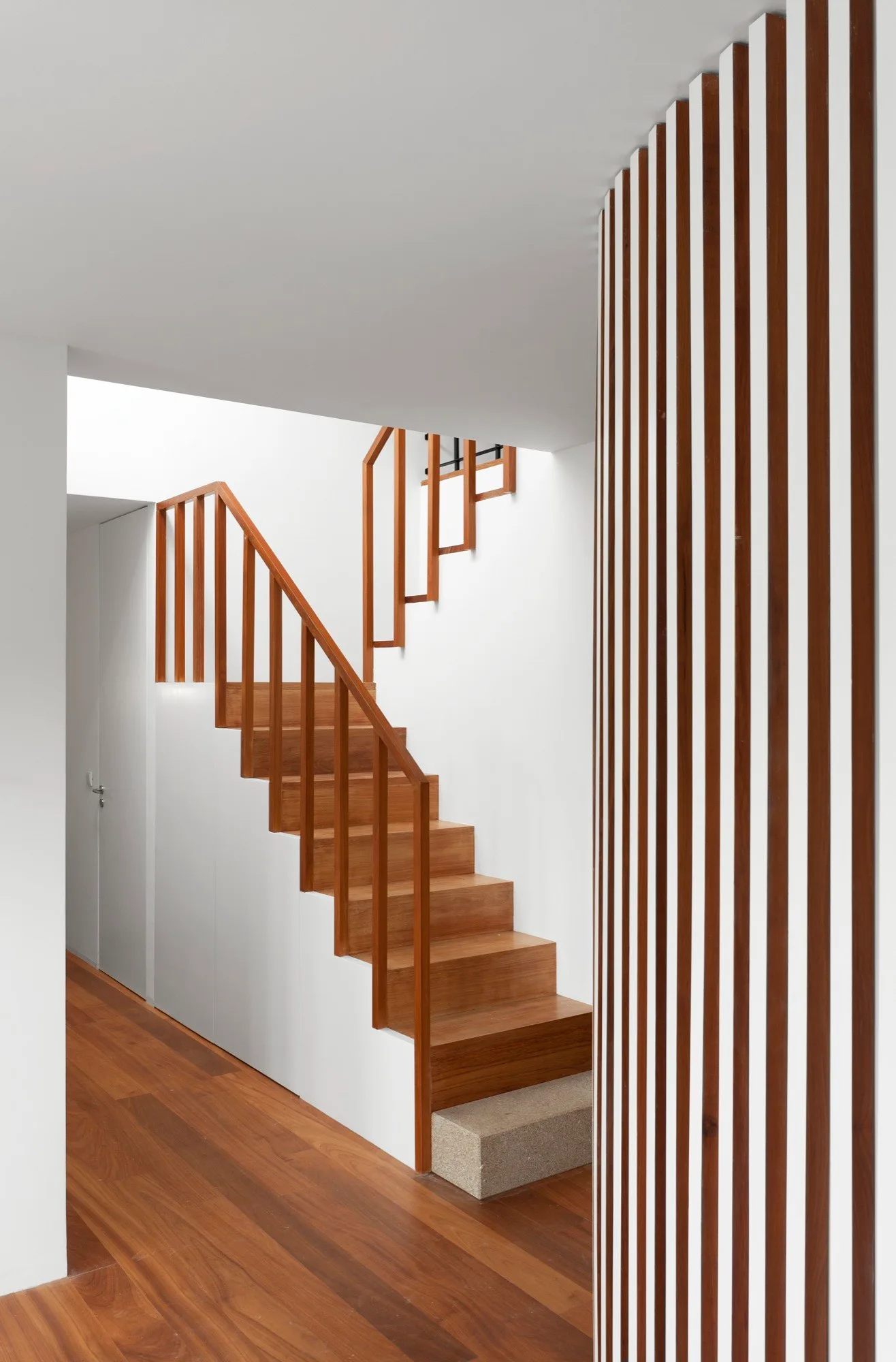 A minimalist wooden staircase with wood treads and a geometric handrail next to a vertical wood slat partition wall.