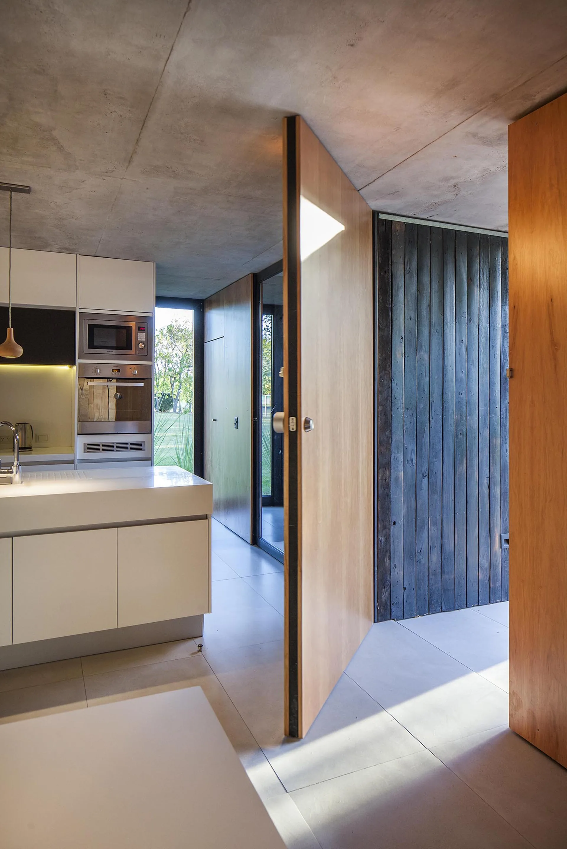 A minimalist kitchen with white cabinetry and a concrete ceiling features a large wooden pivot door opening to an adjacent hallway.