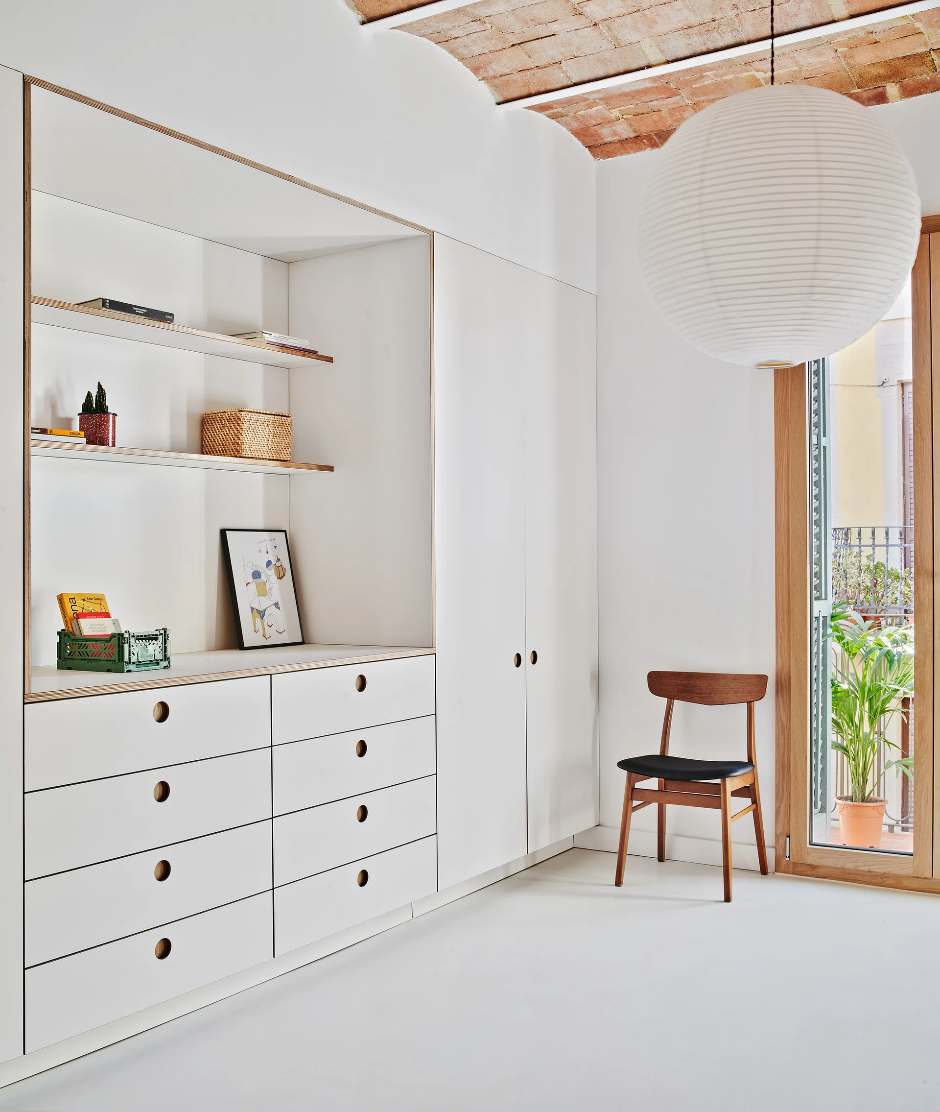A minimalist room featuring white built-in storage cabinets, a wooden chair, and a large paper lantern under an exposed brick ceiling.