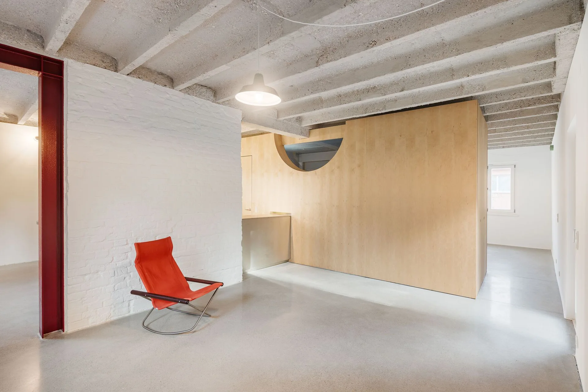 A minimalist space with a red armchair next to a white brick wall, featuring a large plywood partition and exposed concrete ceiling joists.