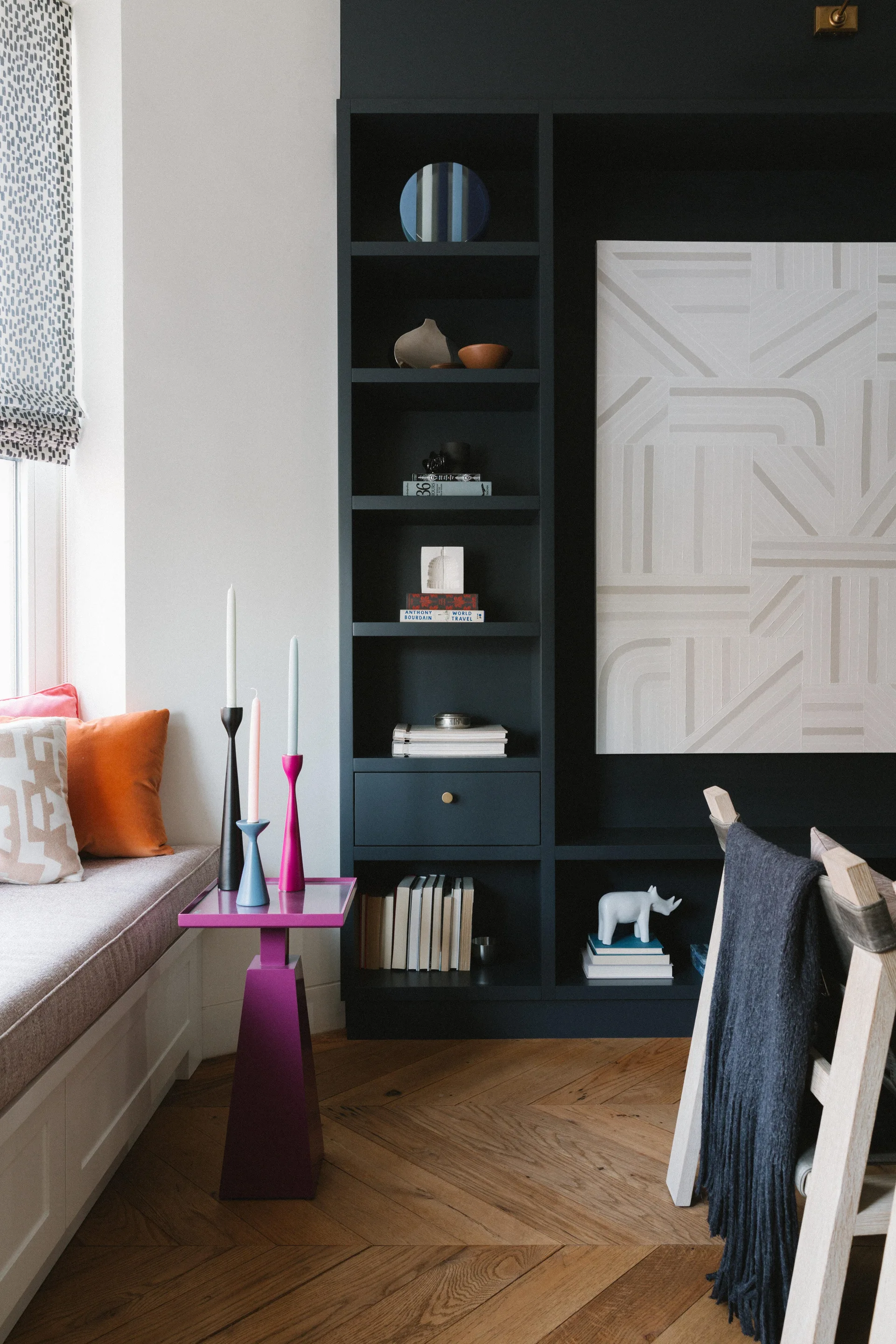 A living room nook shows dark charcoal built-in shelving next to a cushioned window seat, a sculptural magenta side table, and herringbone floors.