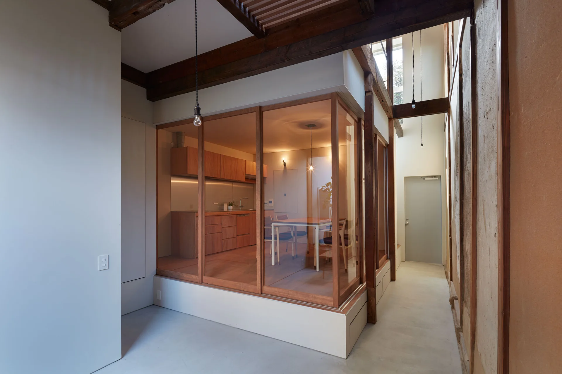 A glass-enclosed kitchen and dining area with wooden cabinetry is set within a room featuring concrete flooring and exposed dark wood ceiling beams.
