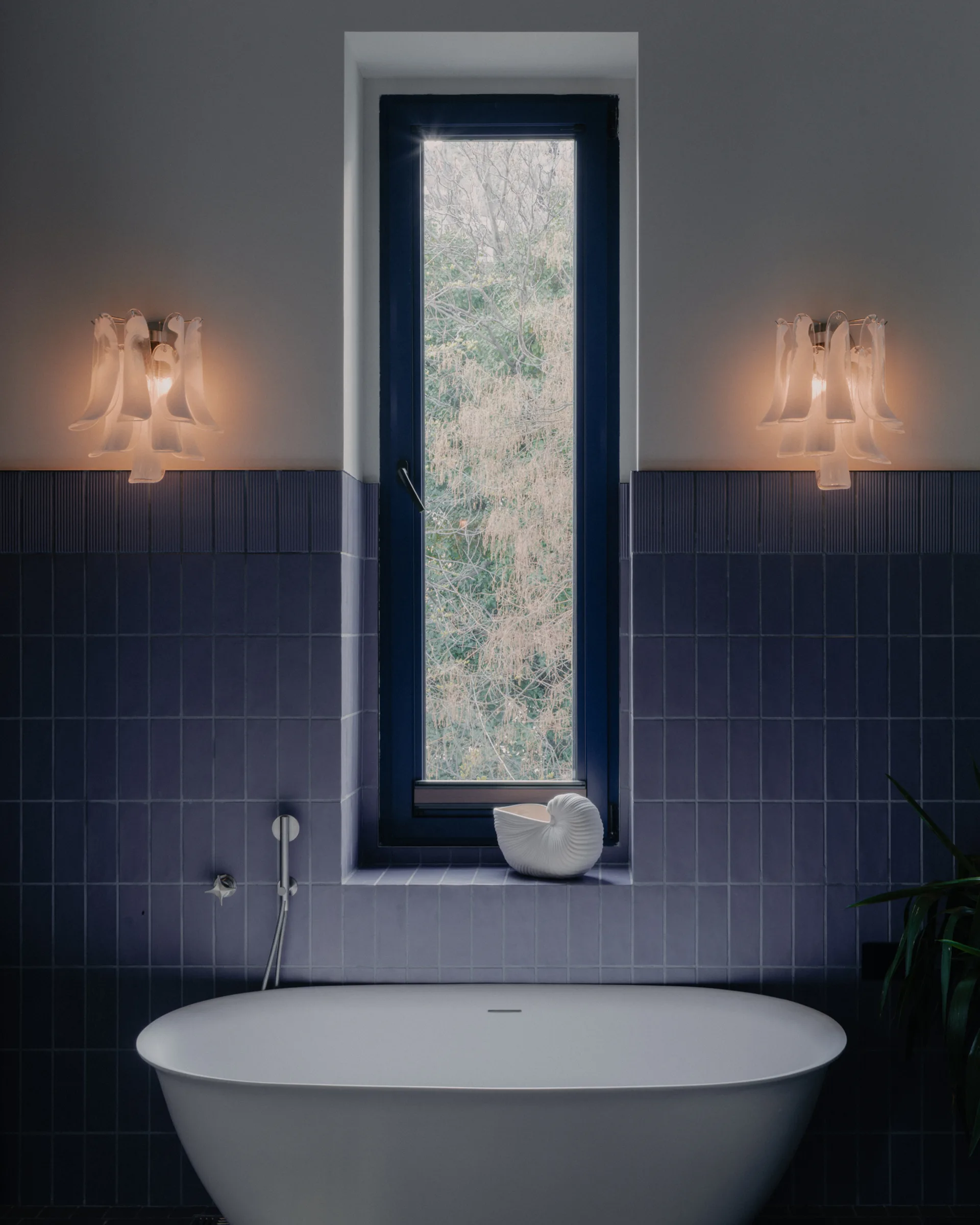 A white freestanding bathtub stands before a narrow window flanked by lit petal-shaped wall sconces against a purple tiled wall.