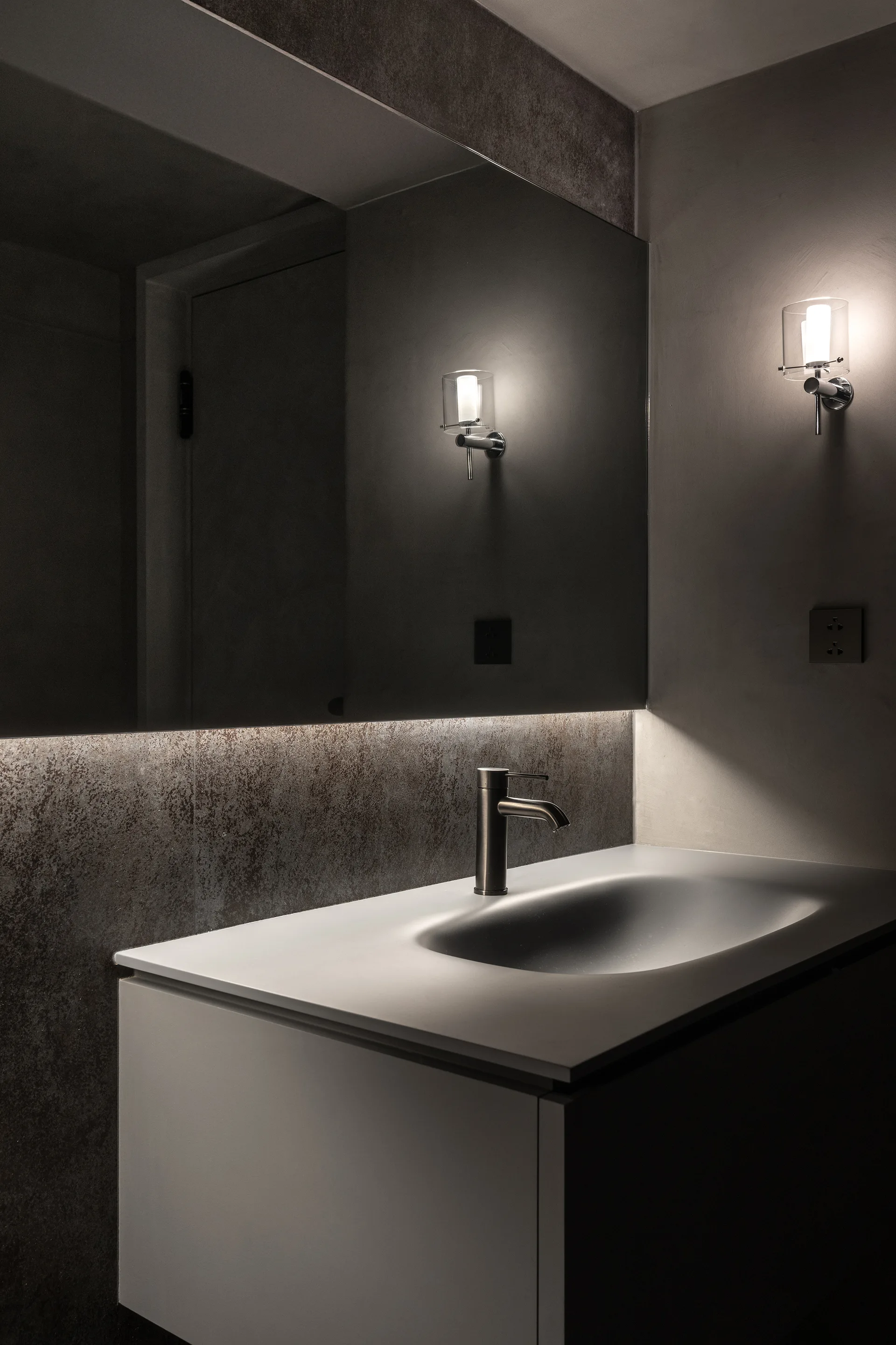 A modern white bathroom vanity with a metal faucet sits against a textured dark grey wall with a large mirror and illuminated glass wall sconces.