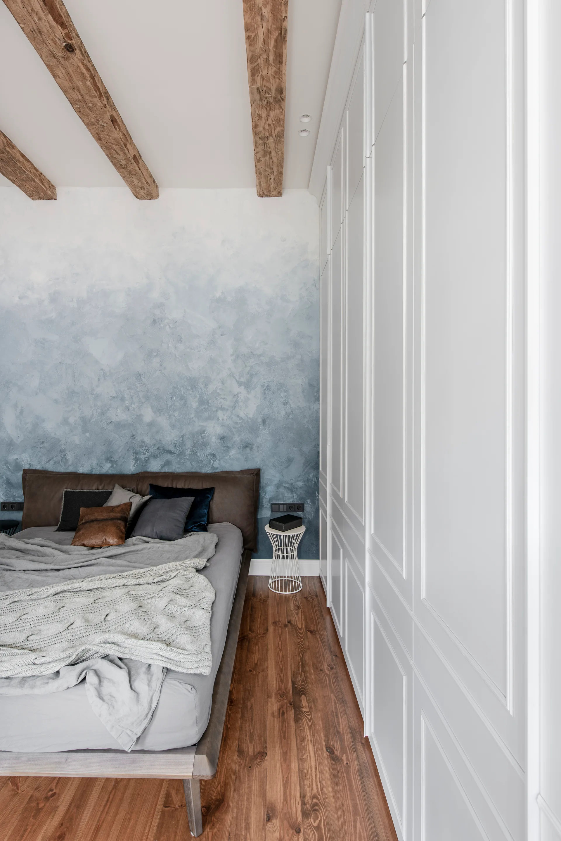 A bedroom features a blue and white ombre accent wall, rustic wood ceiling beams, a leather headboard bed, and a floor-to-ceiling white wardrobe.