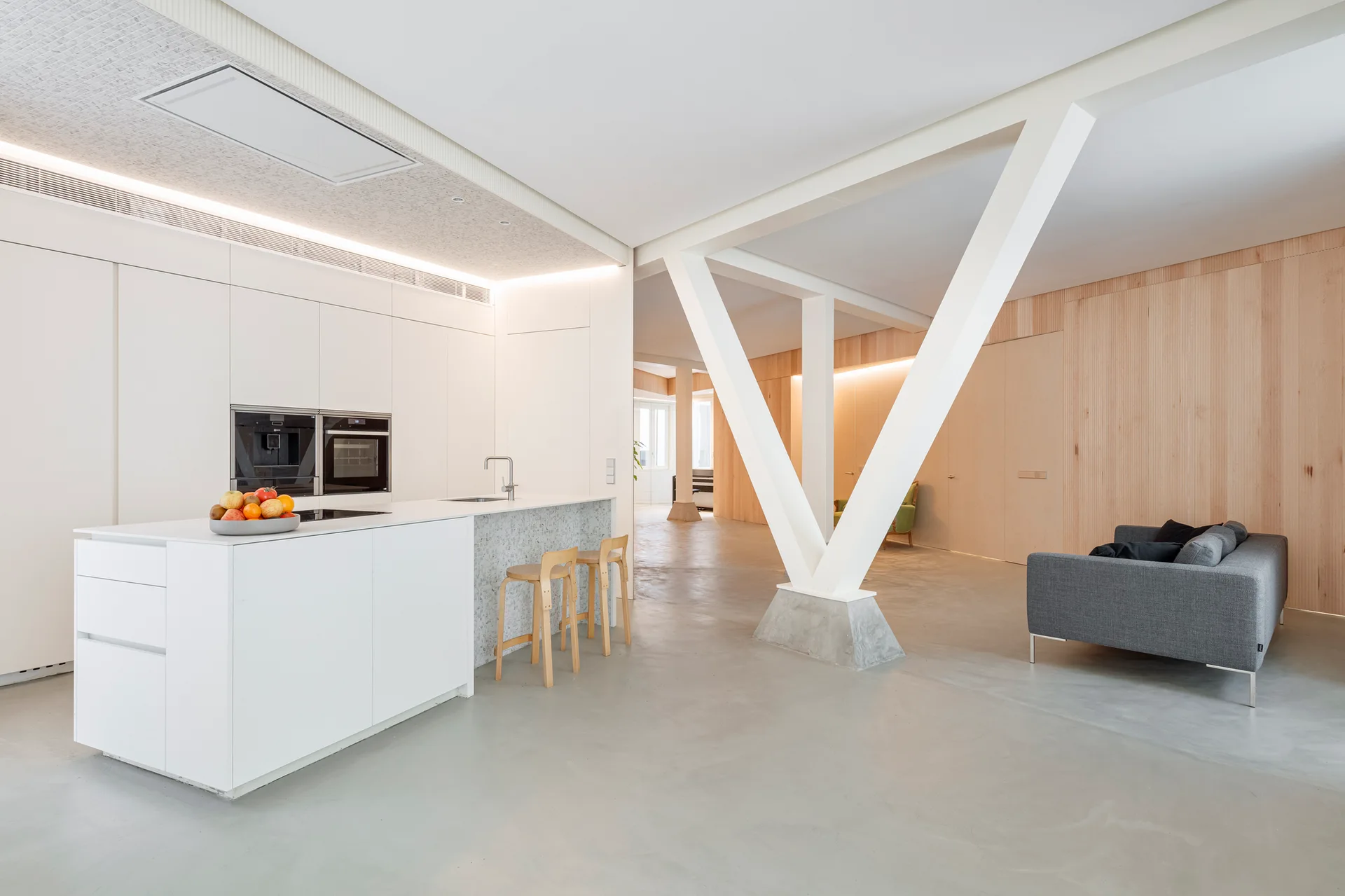 A minimalist open-plan kitchen features a white island with wood stools, a grey sofa, and a prominent white V-shaped structural column.