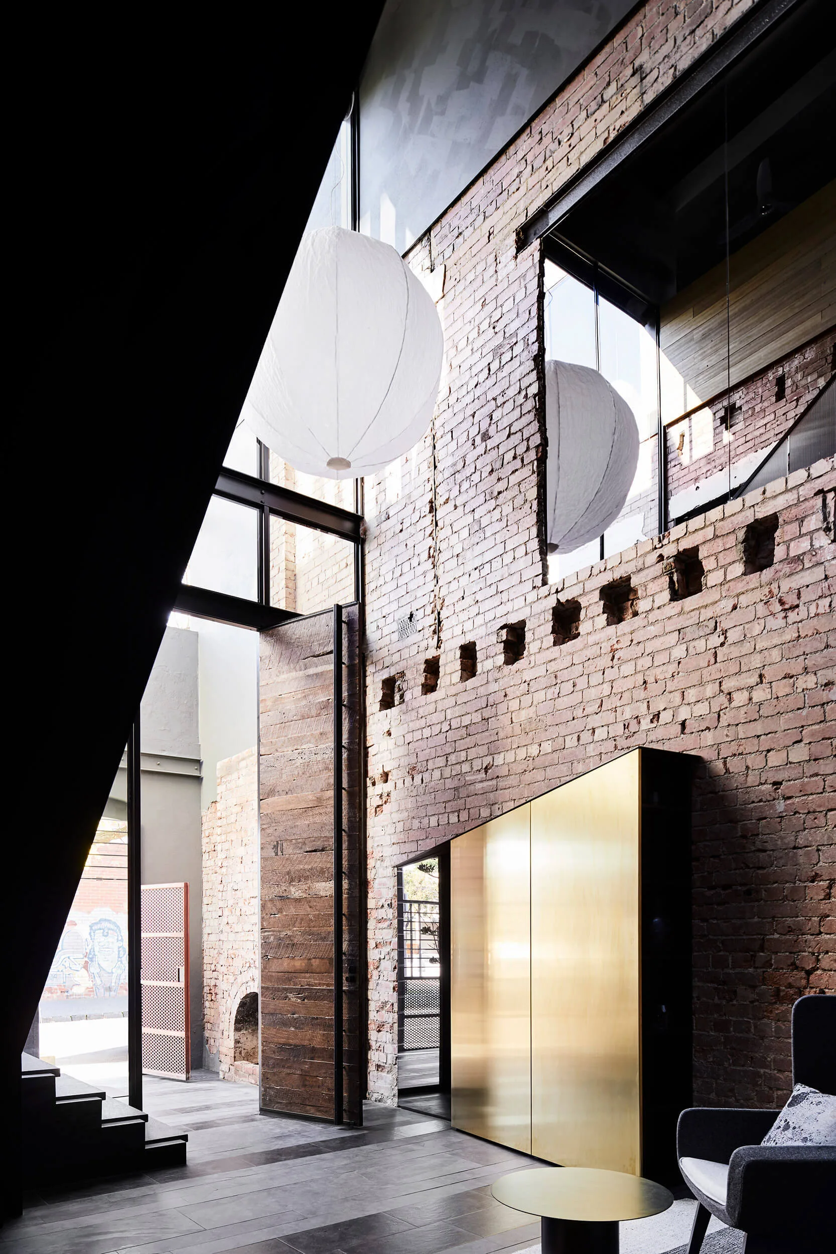 A double-height living room features an exposed brick wall, a large white paper pendant light, and a polished gold storage cabinet on dark tile flooring.