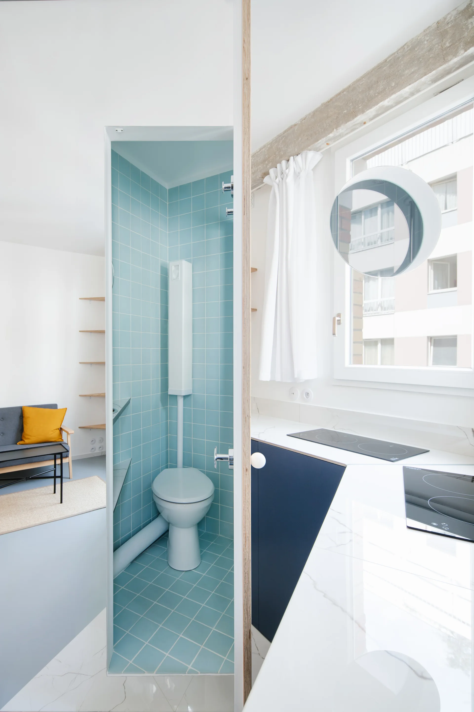An open-concept apartment interior showing a small bathroom with bright blue grid tiles next to a white marble kitchen countertop and a grey sofa.