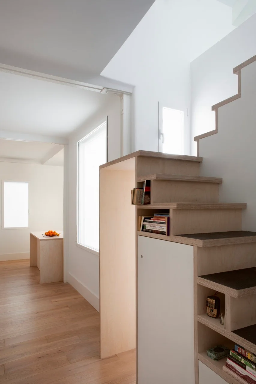 A custom plywood staircase with integrated storage cabinets and open bookshelves stands in a bright room with light wood floors.
