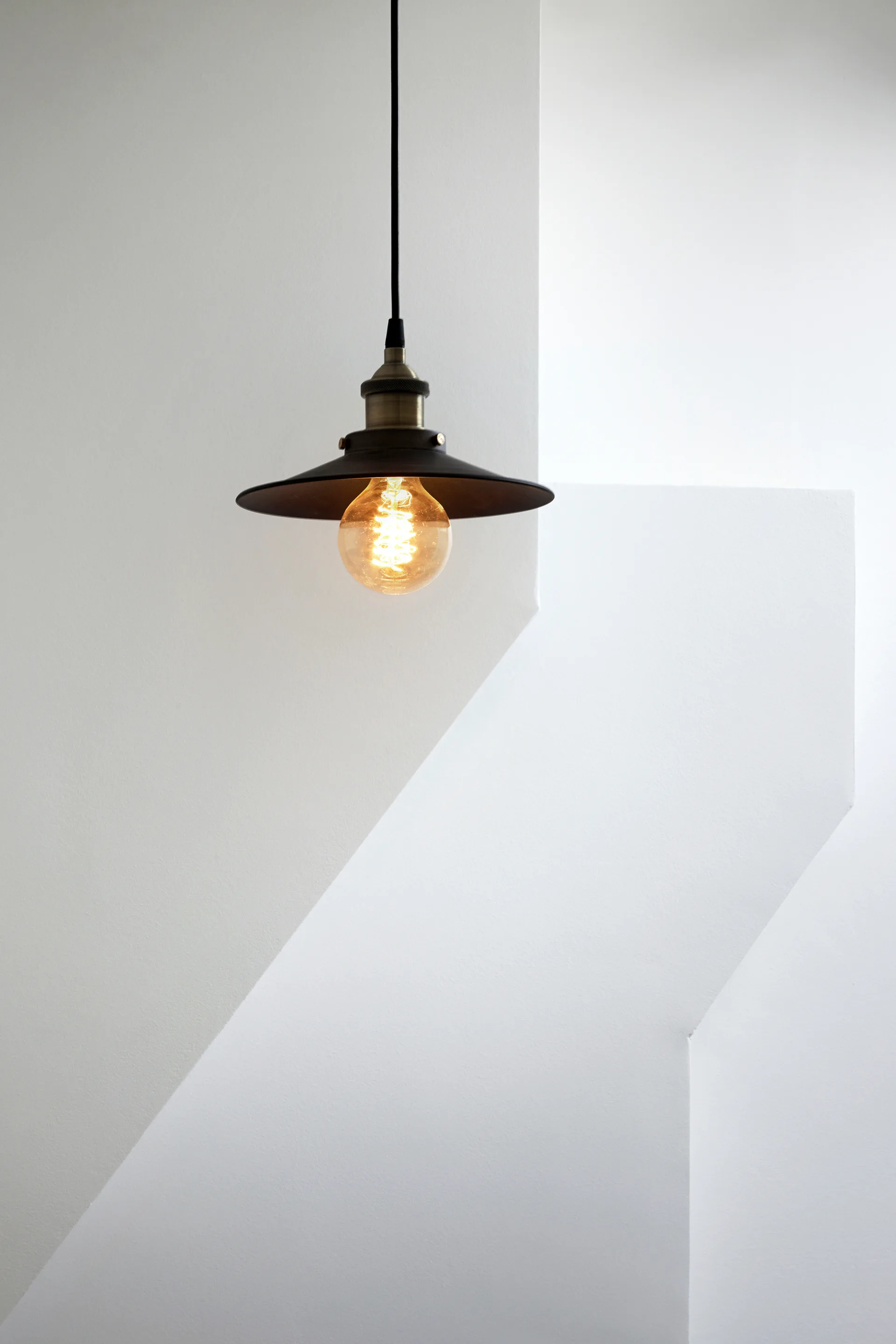 A black industrial-style pendant light with a warm glowing Edison bulb hangs from a black cord in front of a minimalist white geometric wall.