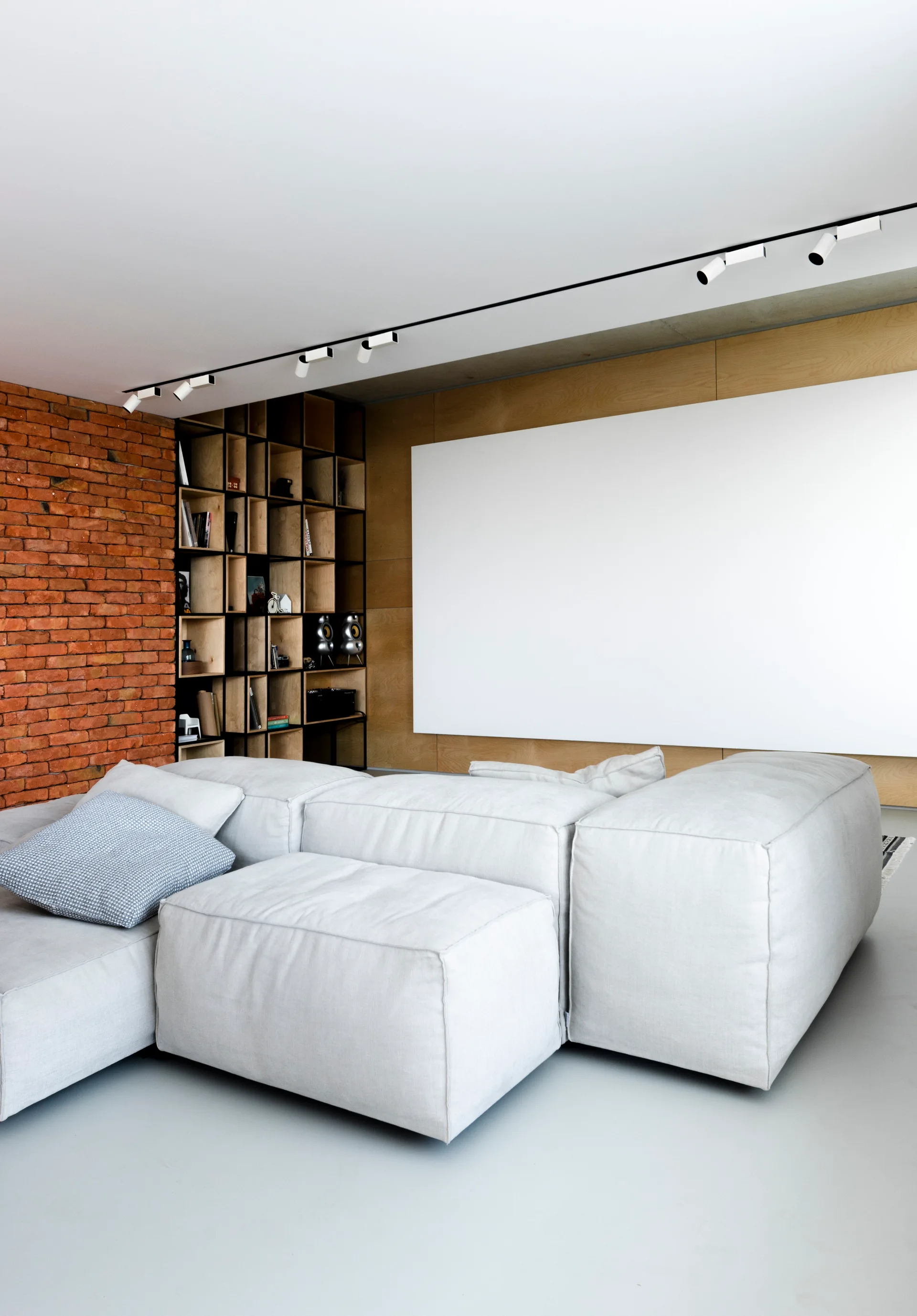 A light grey modular sectional sofa sits in a modern living room featuring an exposed brick wall and a custom wooden grid bookshelf.