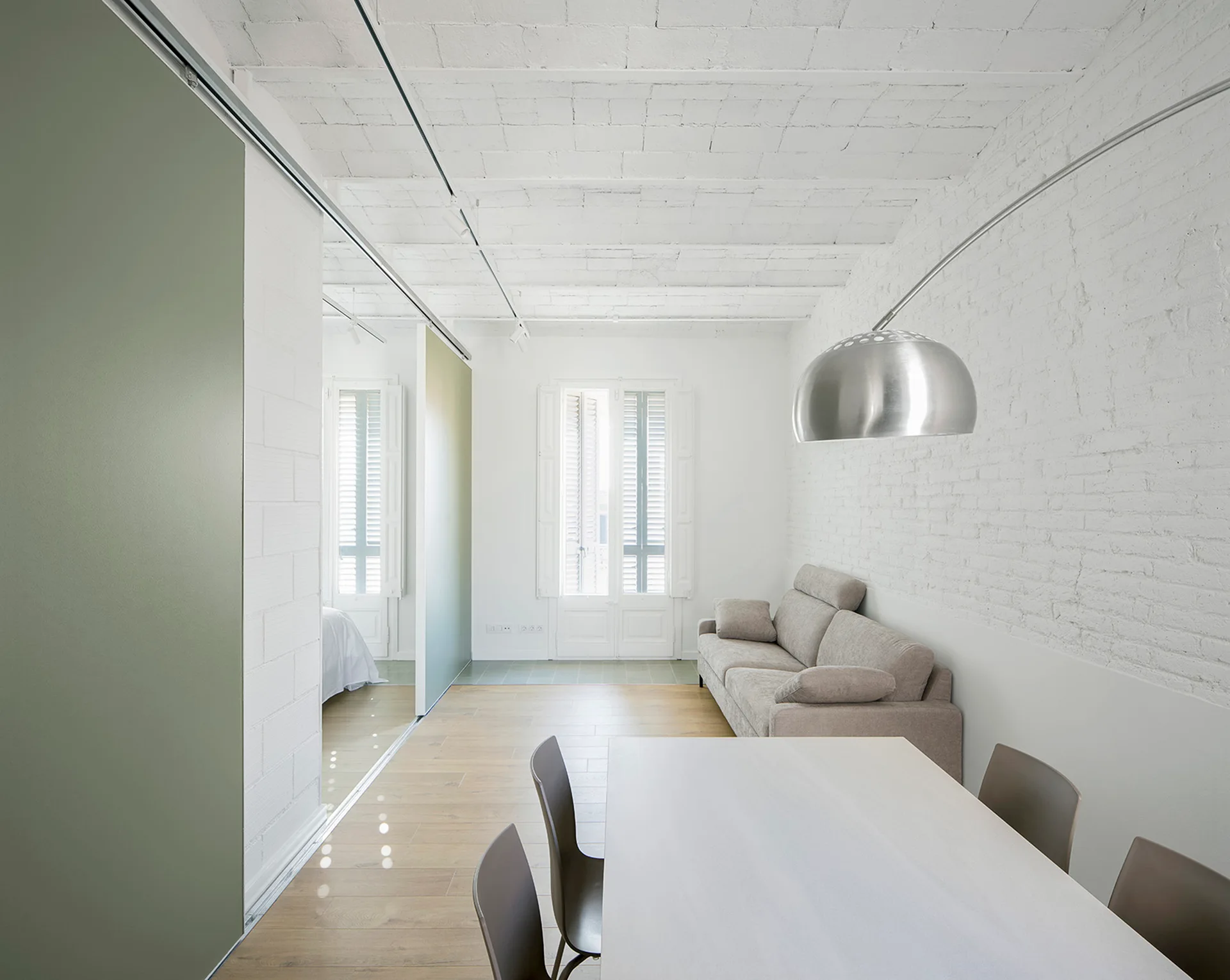 A minimalist living and dining area featuring a white painted brick ceiling, a beige sofa, and large sage green sliding partition walls.