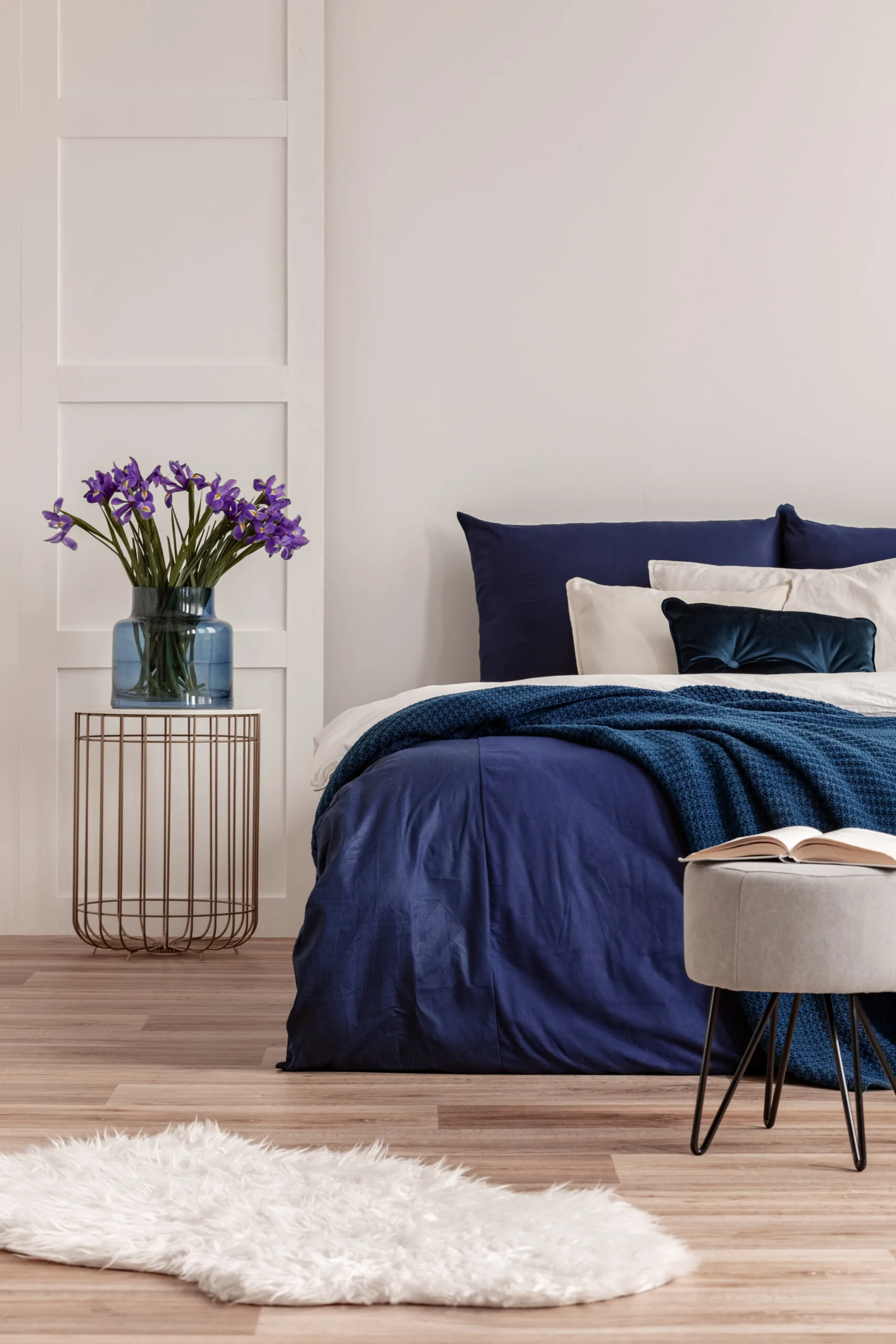 A bedroom features navy blue bedding, a gold wire side table holding a vase of purple flowers, and a small gray ottoman on wood flooring.