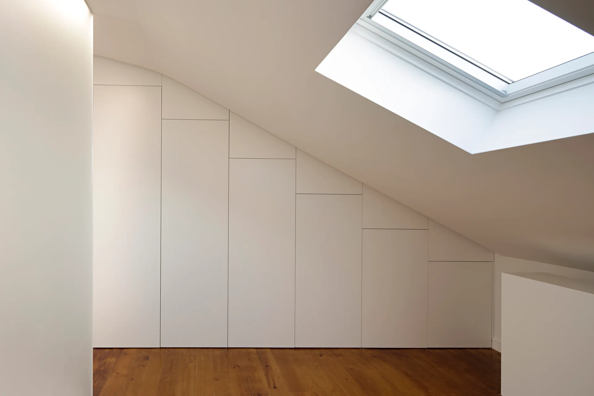 Custom white built-in closets fit beneath a sloped attic ceiling next to a bright skylight and warm wooden floorboards.