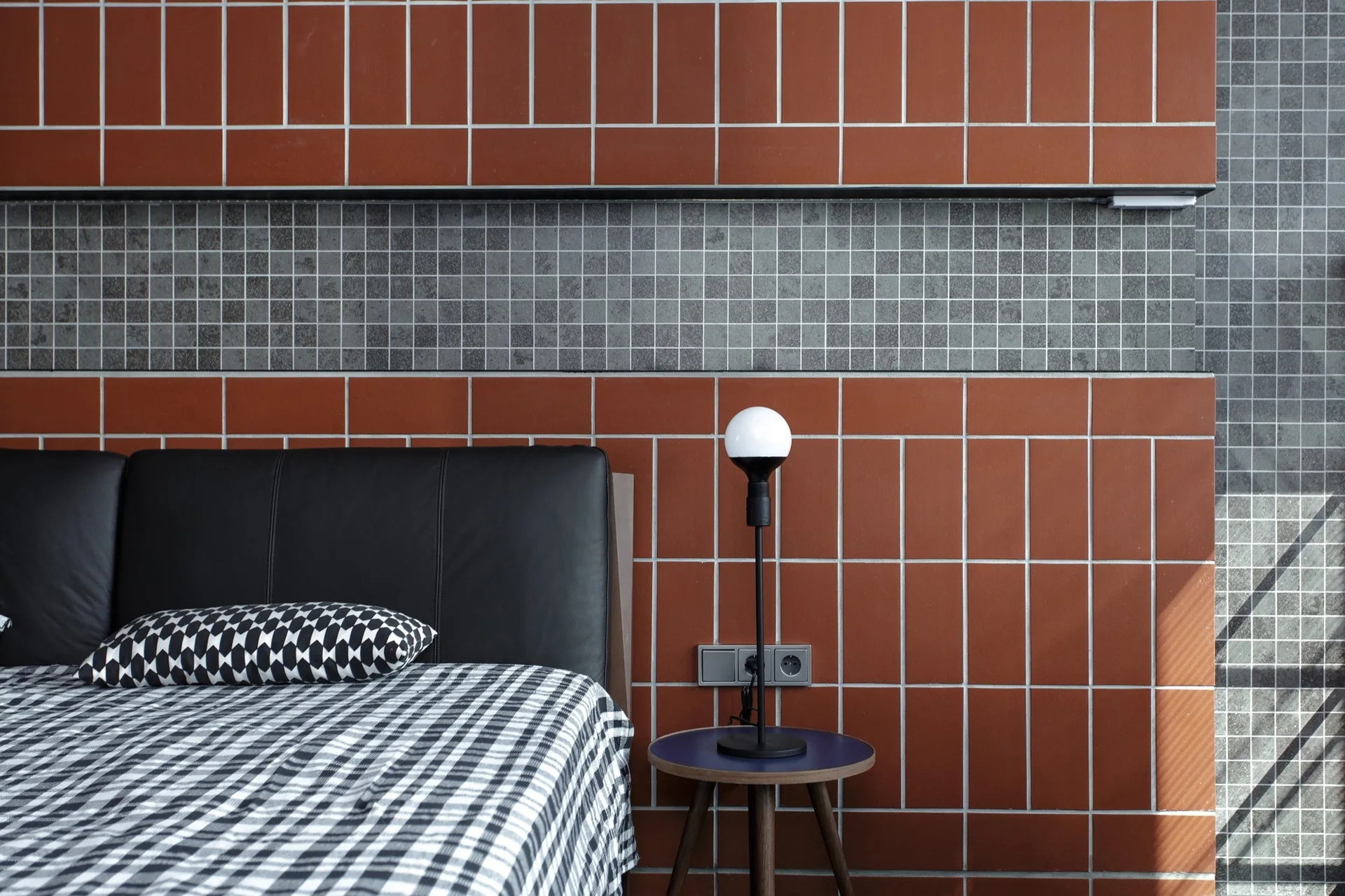 A modern bedroom features a bed with black and white checked bedding next to a small wooden nightstand against a rust red and grey tiled accent wall.