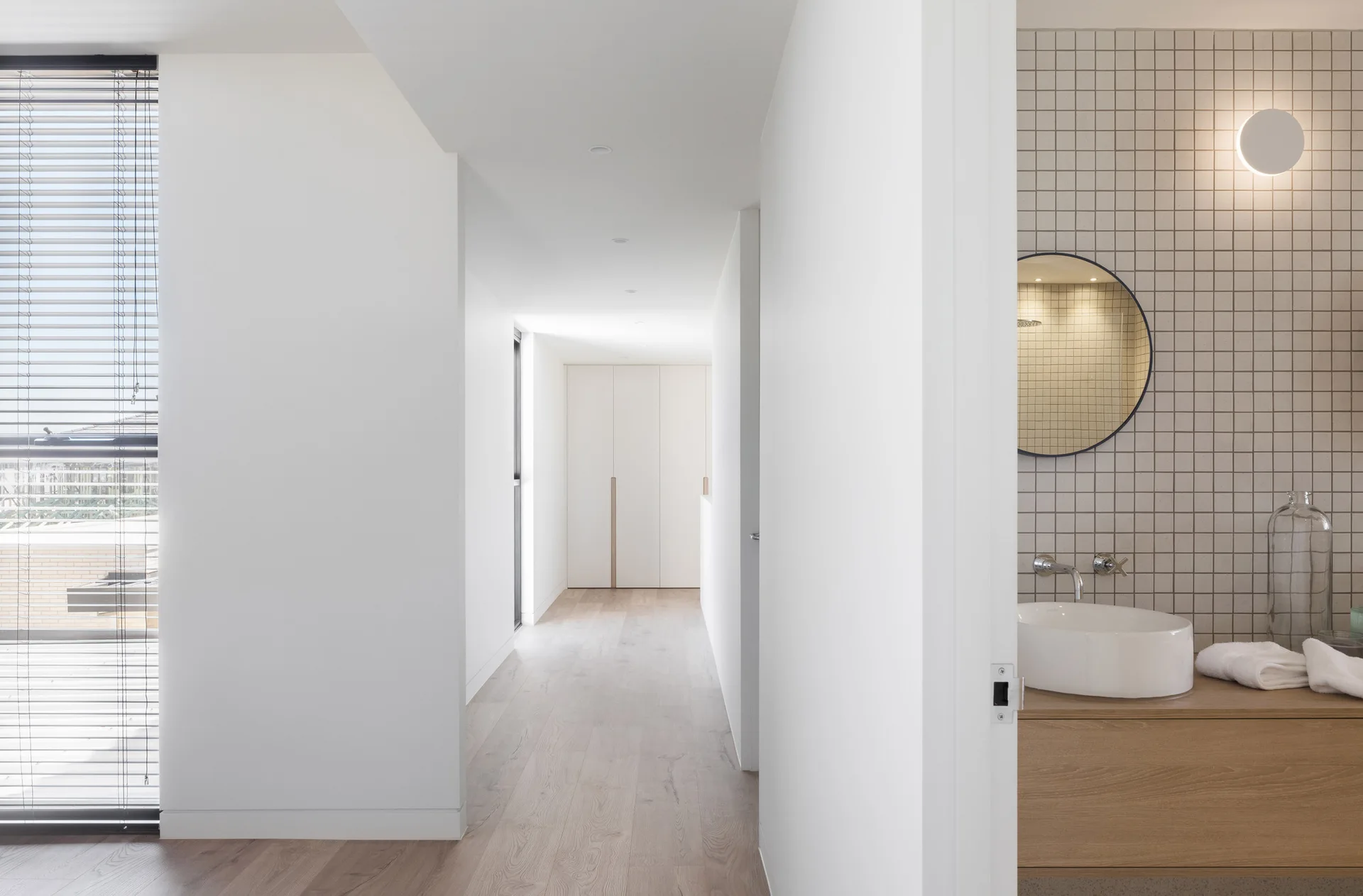 A bright white hallway leads to a modern bathroom featuring a light wood vanity, white vessel sink, and a square-tiled wall with a round mirror.