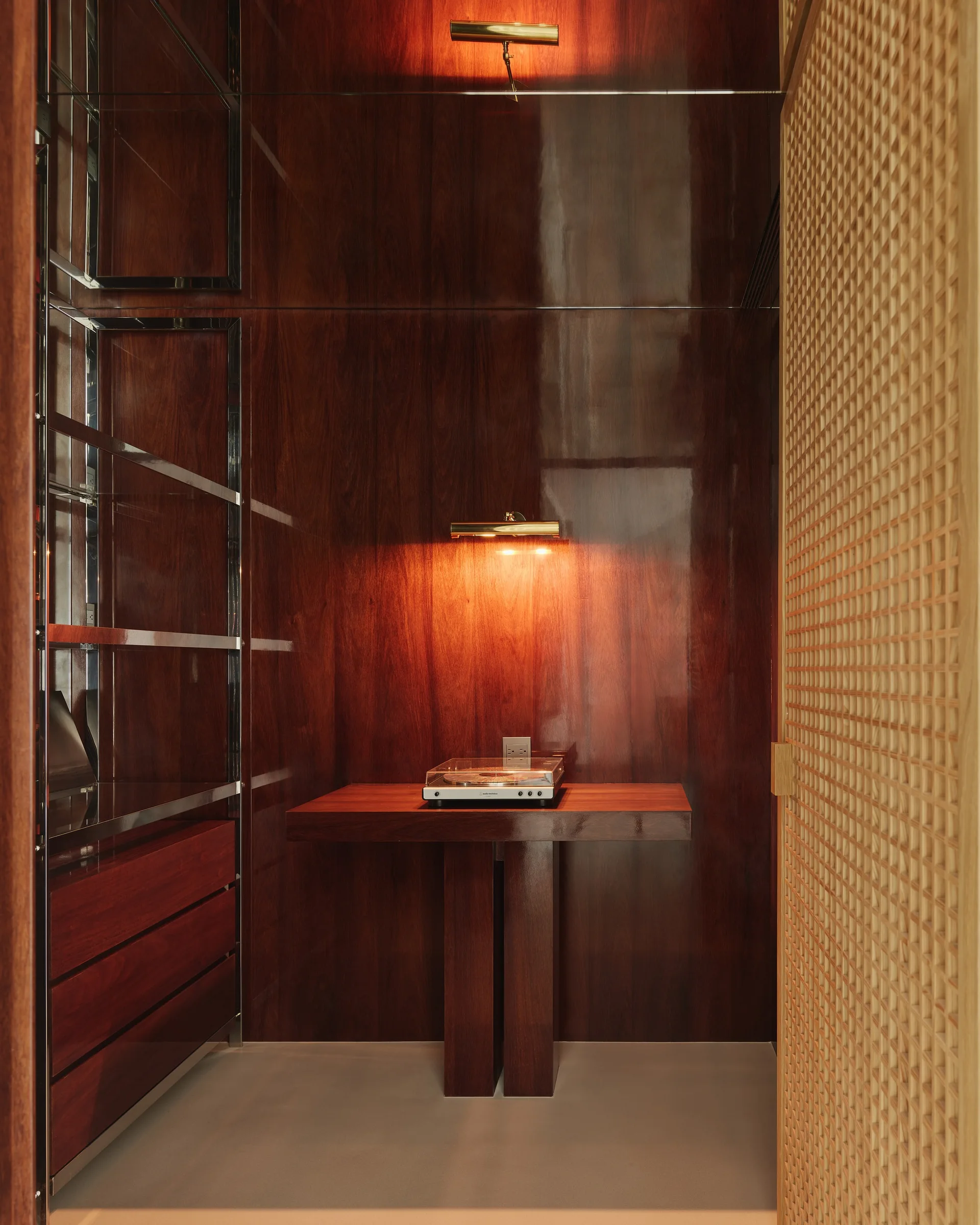 A dark wood paneled niche features a central wood console with a turntable, illuminated by a warm brass picture light, next to a light wood lattice door.