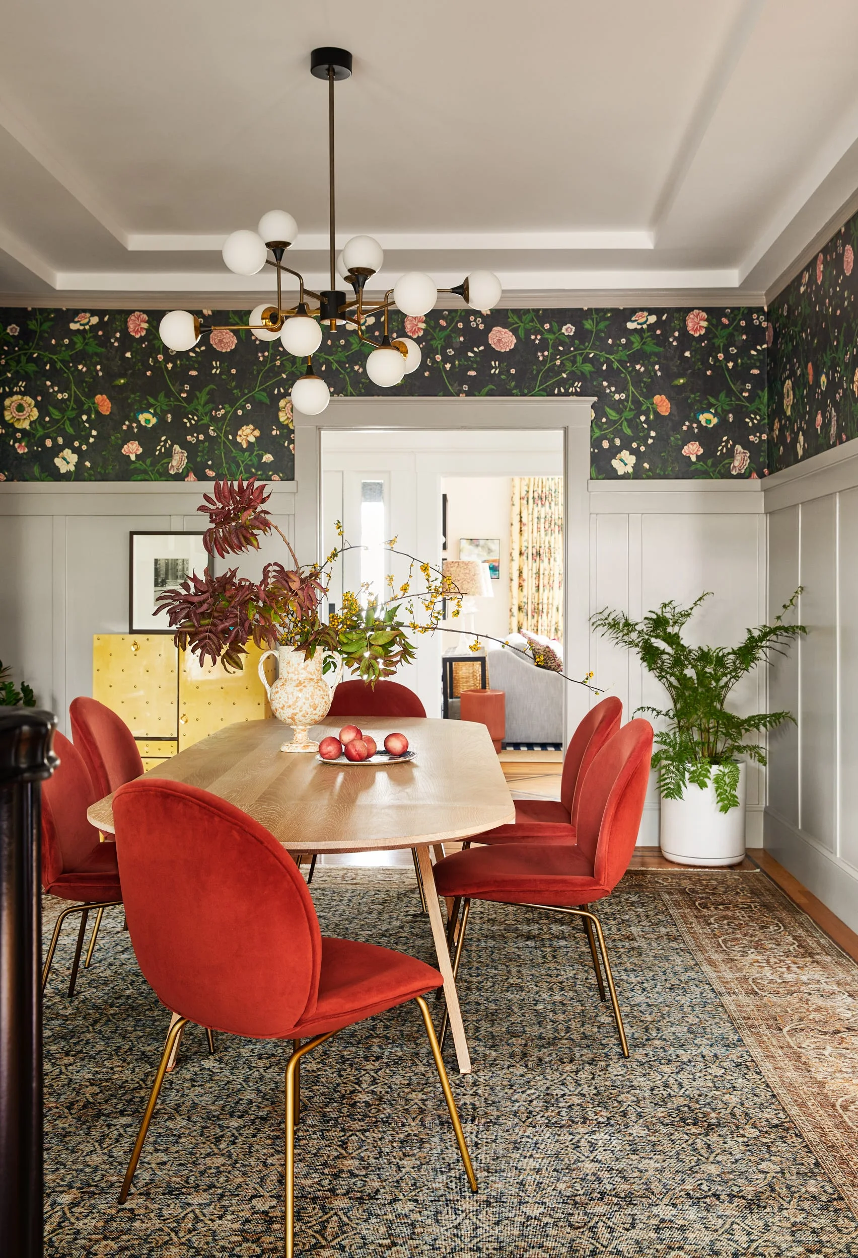A dining room features a light wood table and red velvet chairs beneath a globe chandelier with dark floral wallpaper and gray wainscoting.