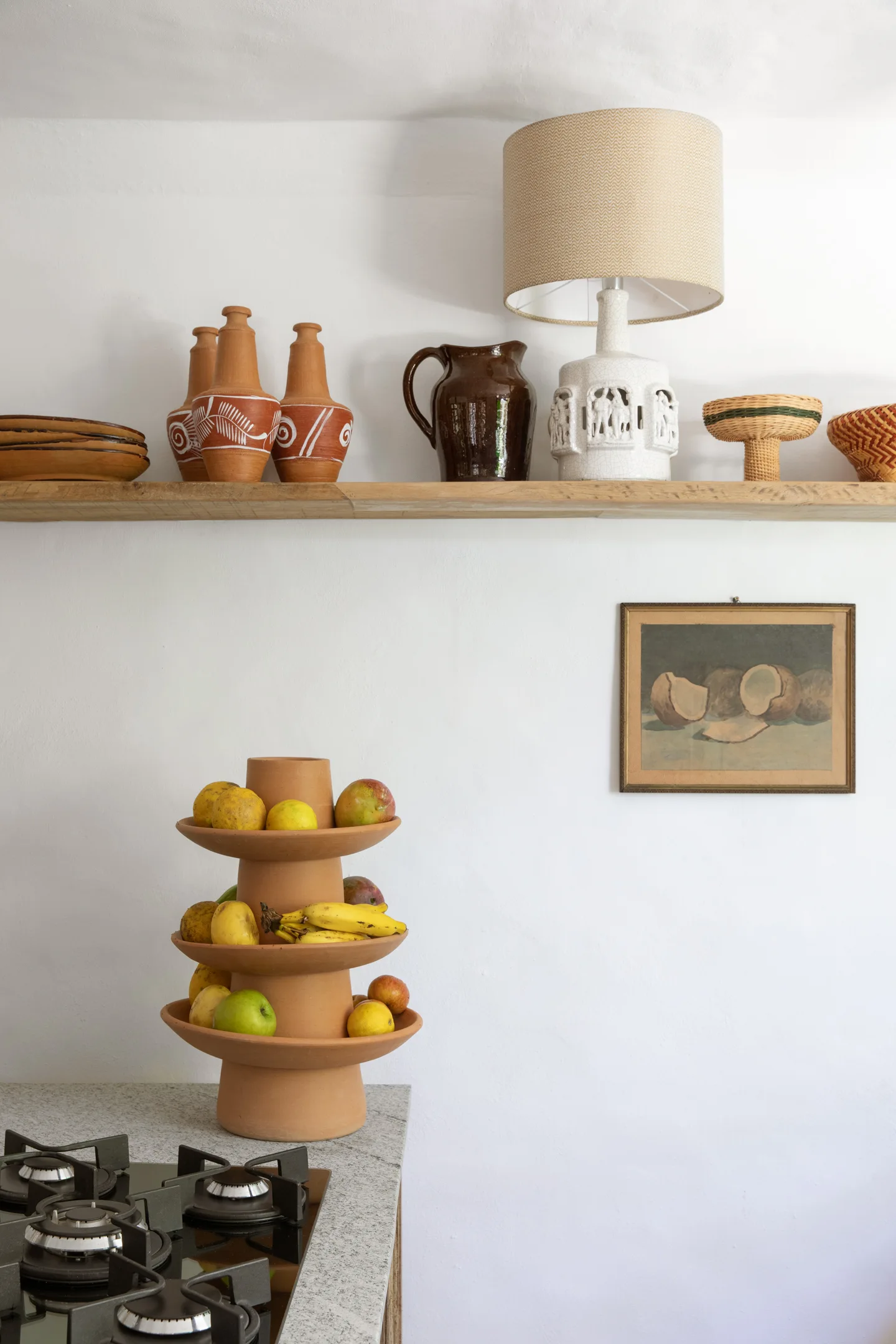 A wooden floating shelf with terracotta pots and a lamp sits above a kitchen counter with a tiered ceramic fruit stand.