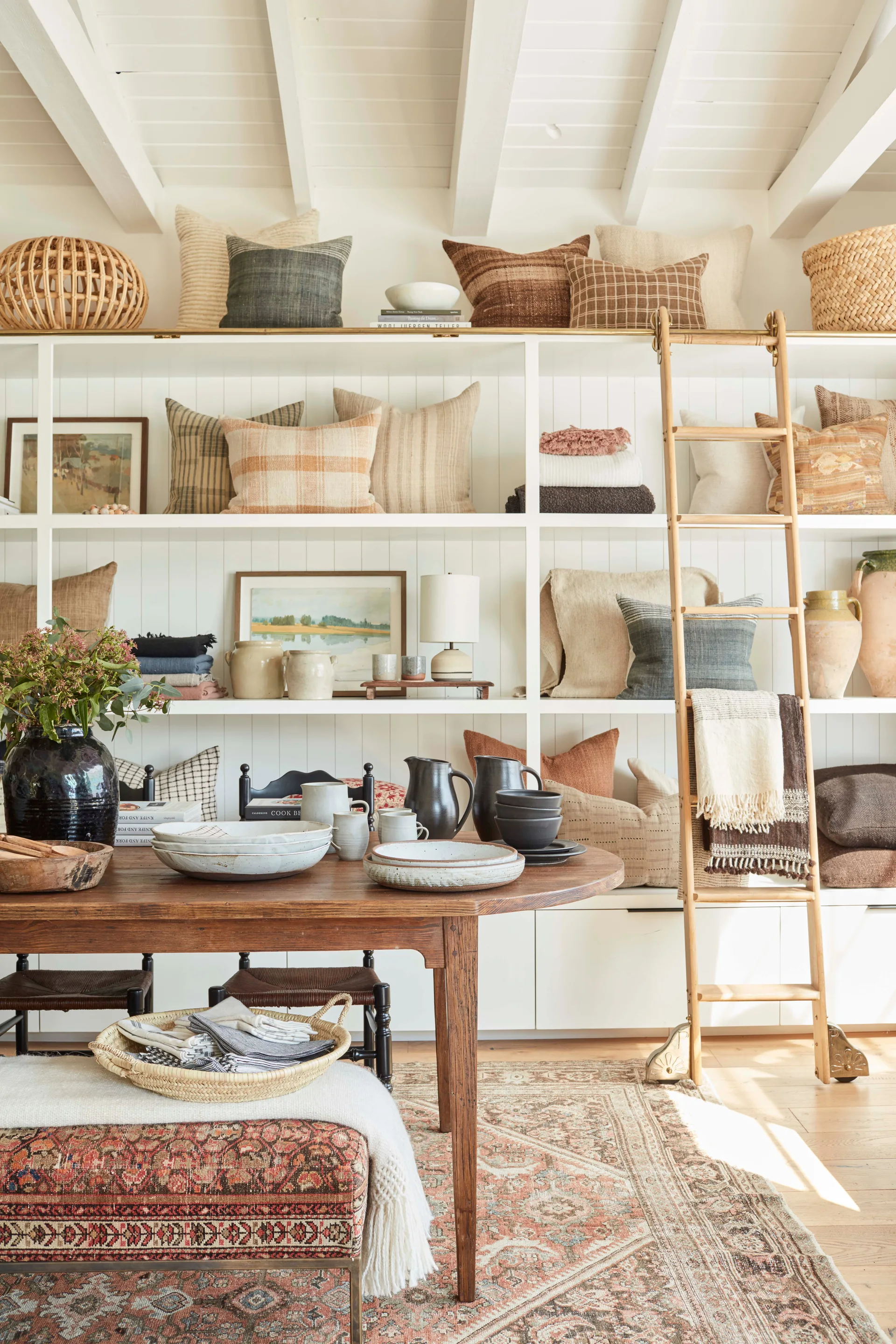 A wooden dining table sits in front of a large white built-in shelving unit filled with patterned pillows and a rolling wooden ladder.