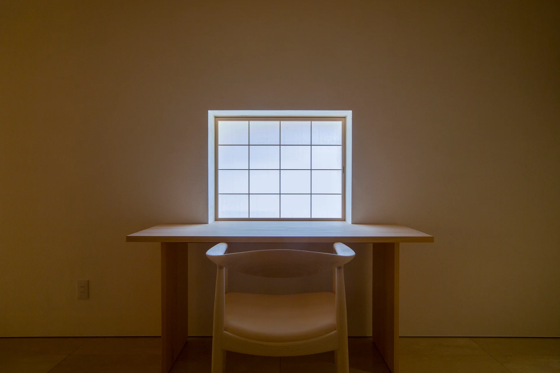 A light wood desk and curved matching chair are centered beneath a square shoji-style grid window on a smooth beige wall.
