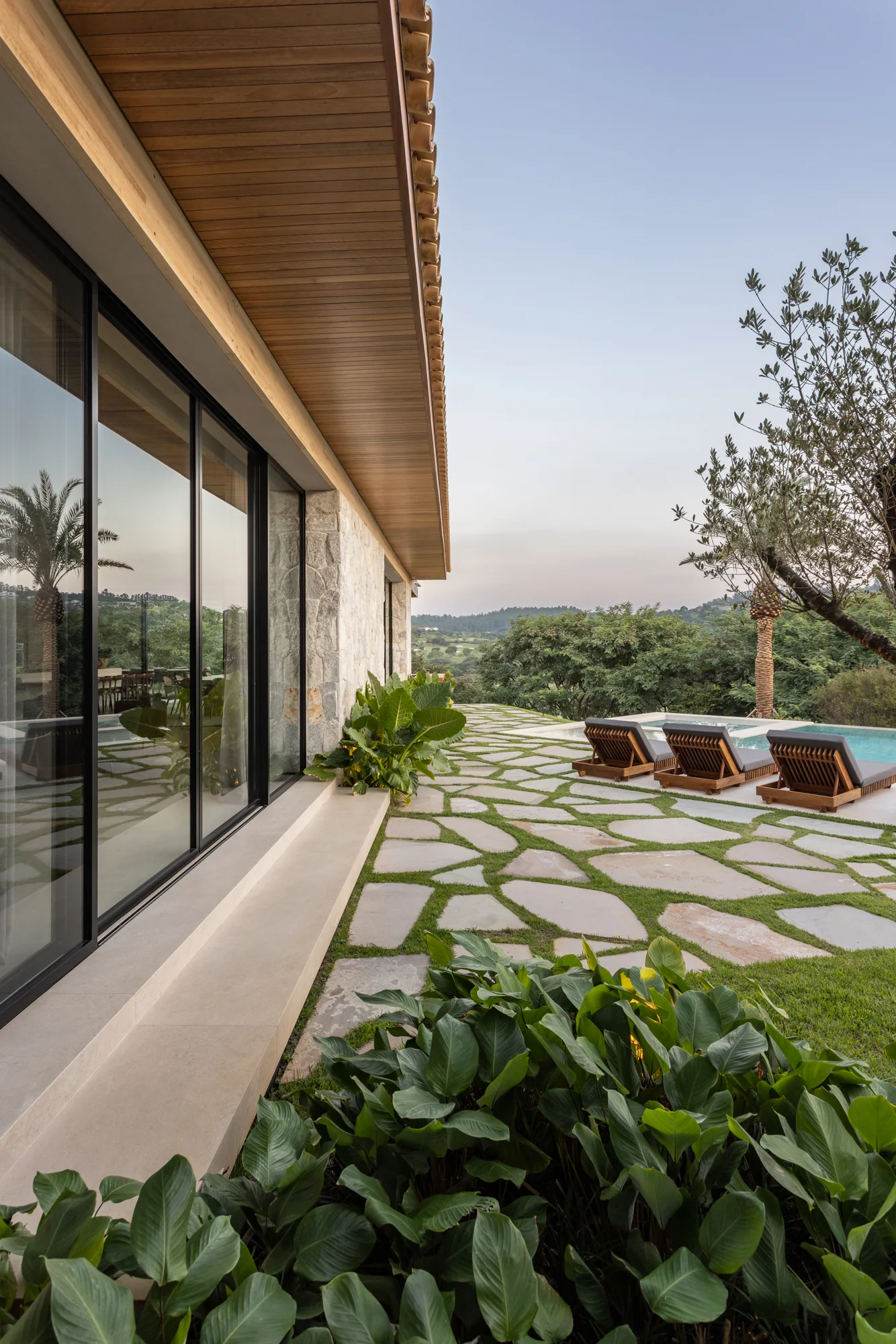 A modern organic patio with irregular flagstone paving and grass joints features wooden lounge chairs next to a swimming pool under a wood soffit.