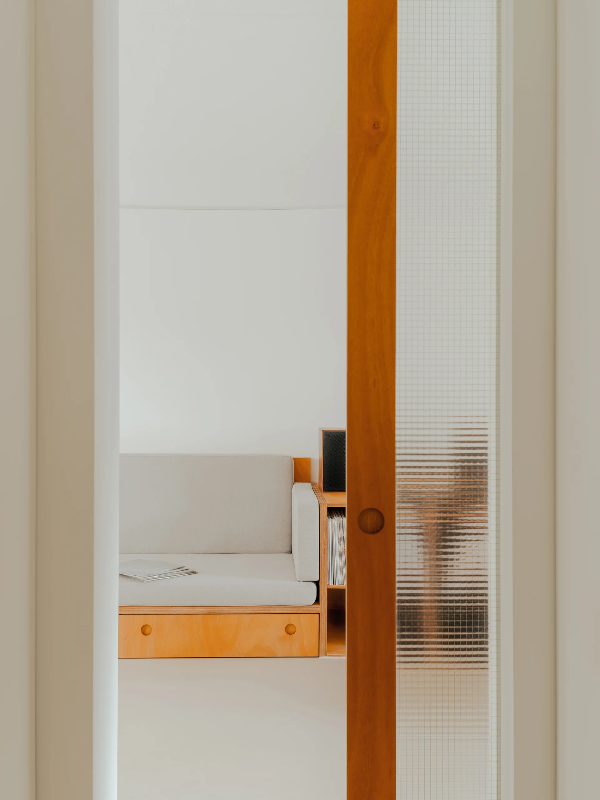 A minimalist light gray sofa with a plywood frame is viewed through a sliding doorway featuring a reeded glass panel.