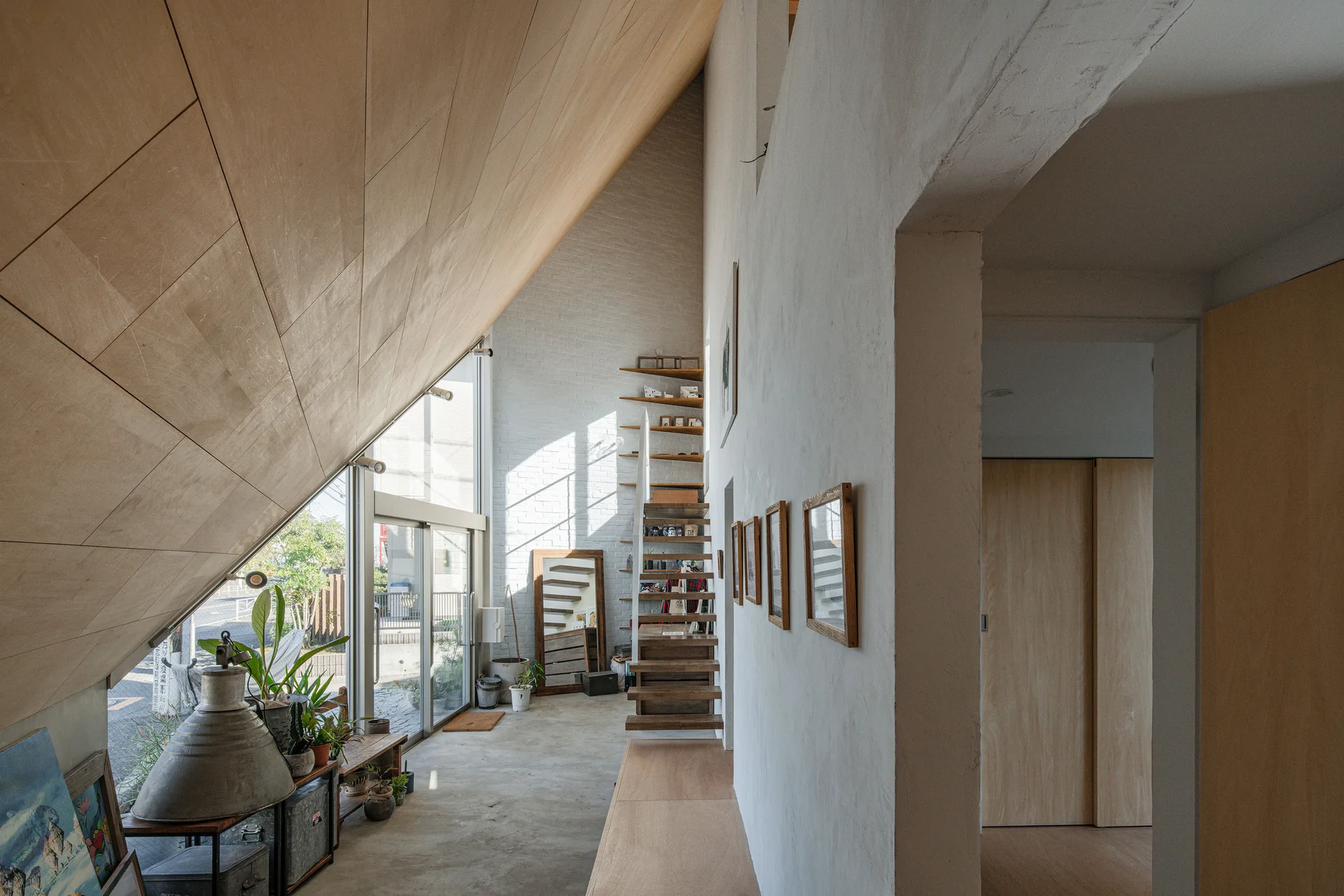 A bright living area features a slanted wood ceiling, a floating wooden staircase against a white brick wall, and smooth concrete flooring.