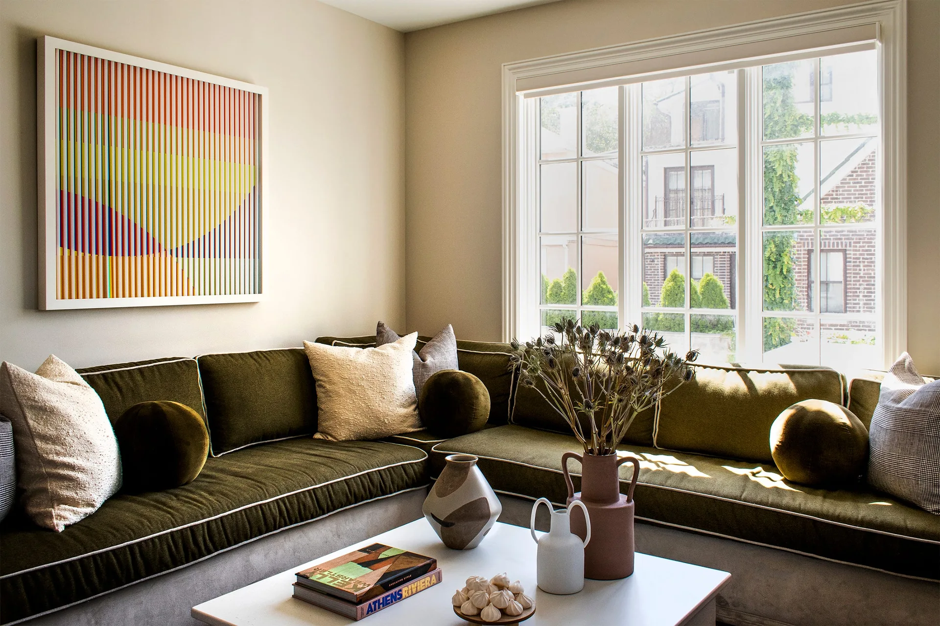 An olive green sectional sofa with white piping and round bolster pillows sits in a living room corner next to a large multi-pane window.