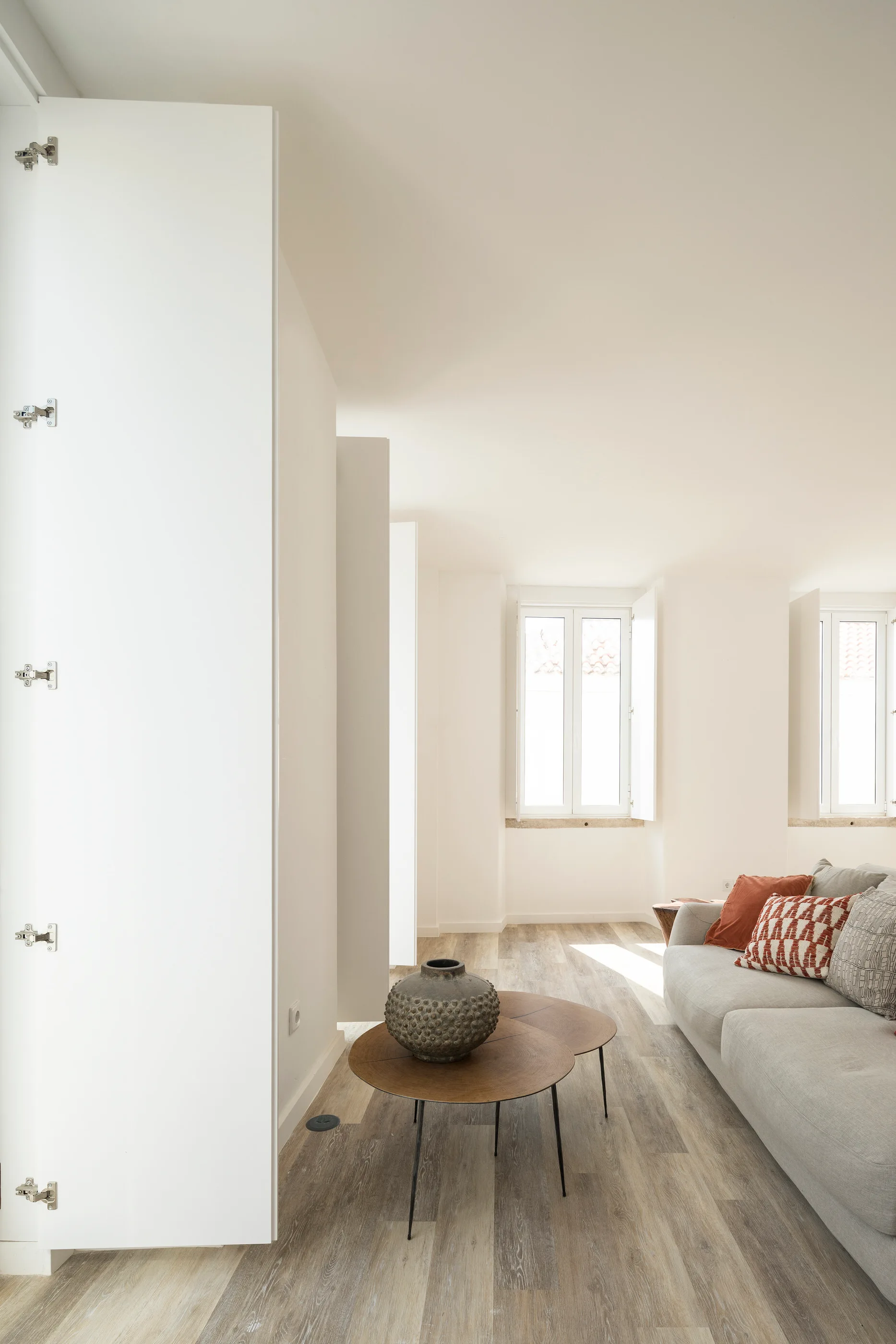 A minimalist living room features a grey sofa with patterned pillows, nested round wood coffee tables, and built-in white cabinets.