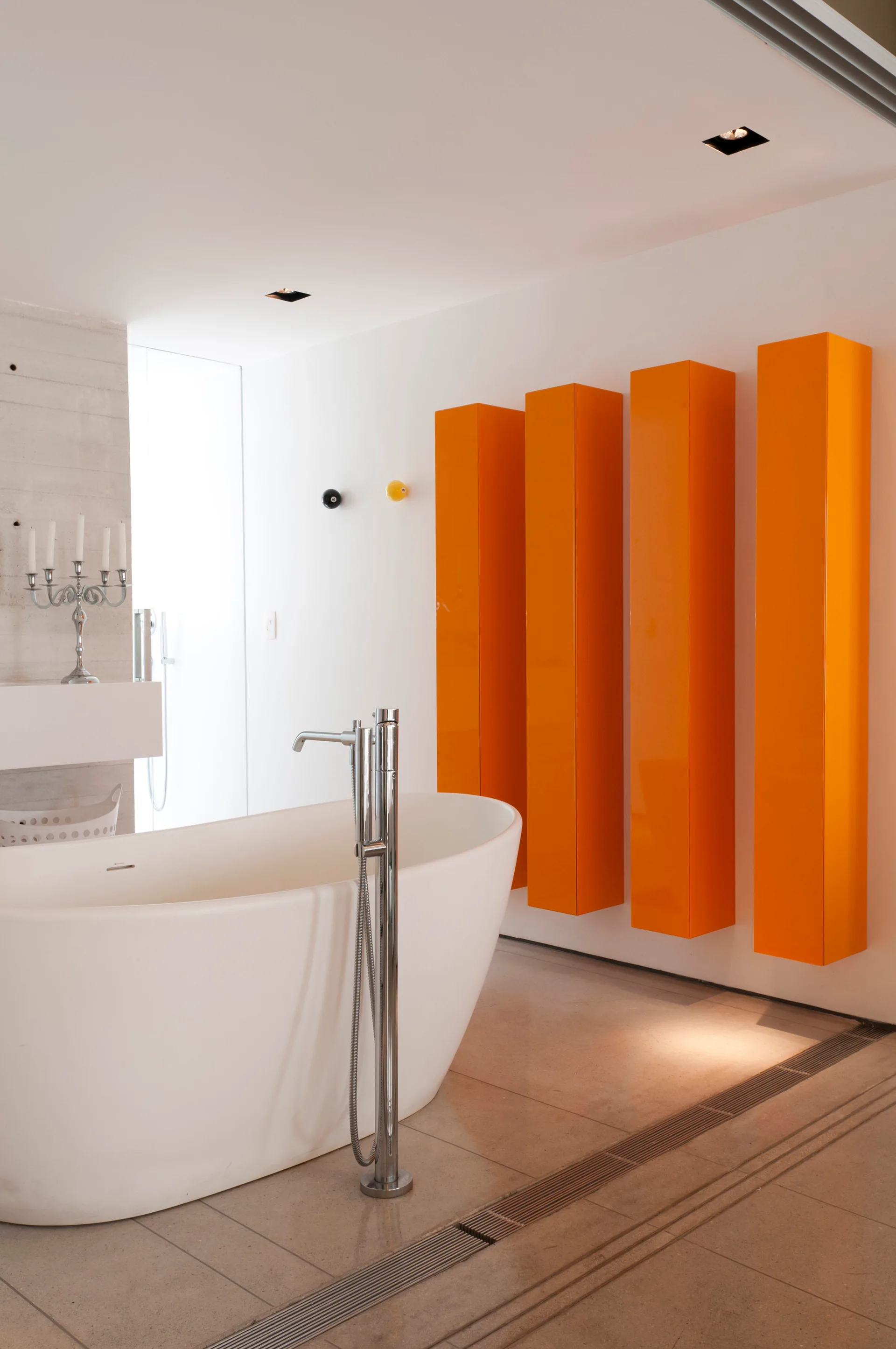 A minimalist white bathroom features a freestanding tub with a tall chrome faucet next to four bright orange vertical cabinets mounted on the wall.