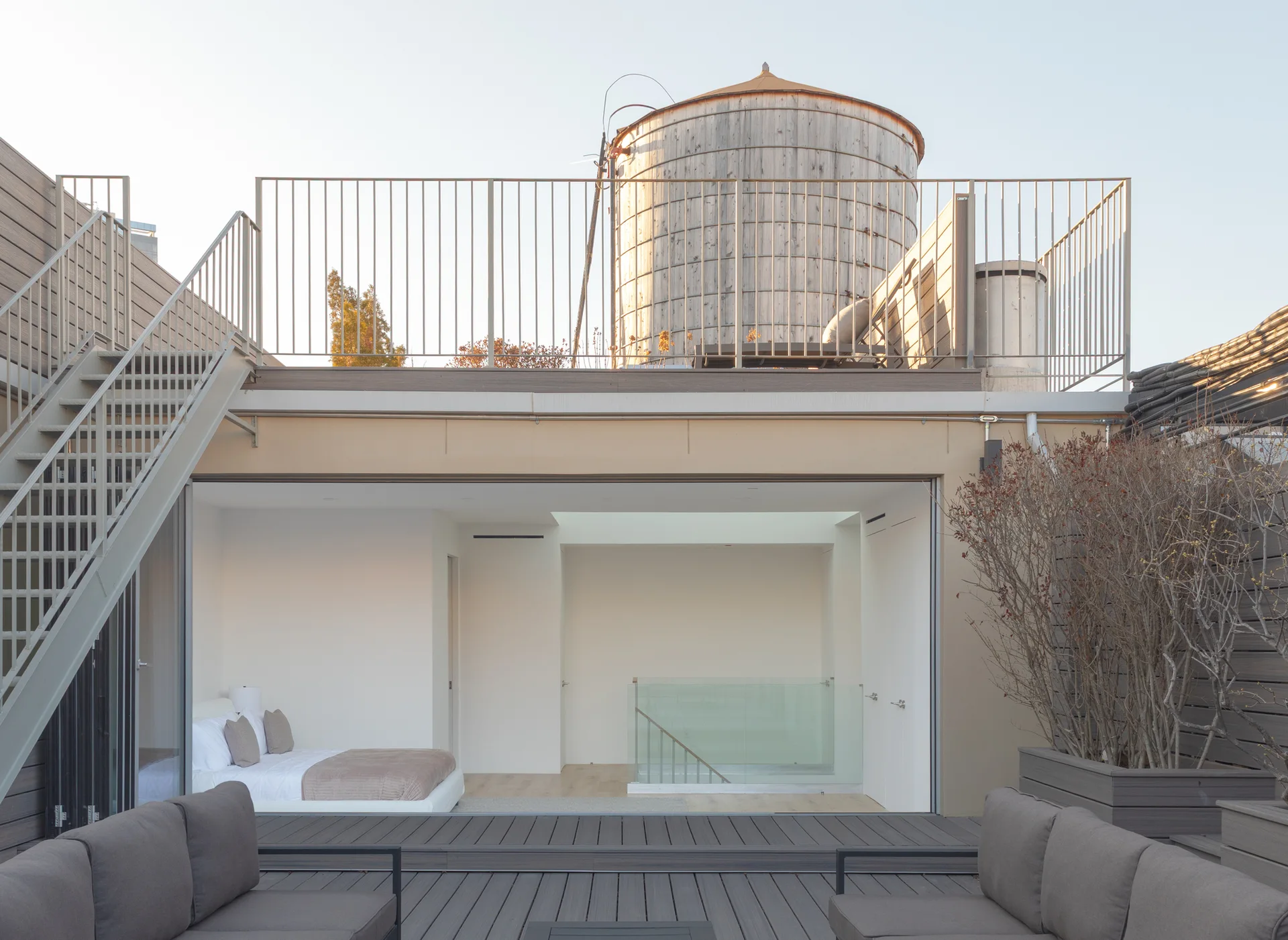 A modern rooftop terrace with gray composite decking and outdoor sofas looks into an adjacent bedroom with a white bed and minimalist interior.