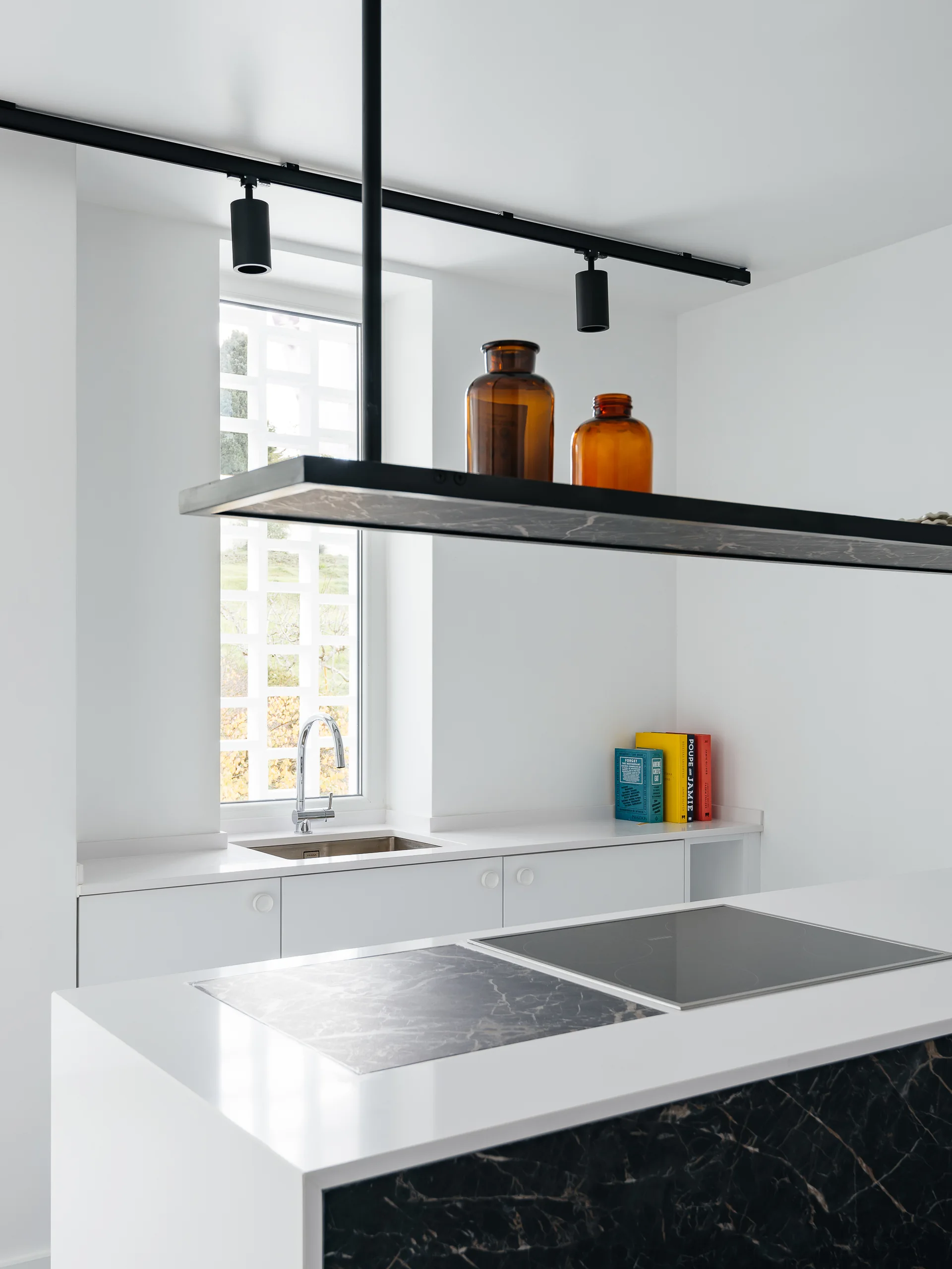 A minimalist white kitchen featuring a central island with a dark marble inlay, a suspended black metal shelf, and base cabinetry beneath a window.