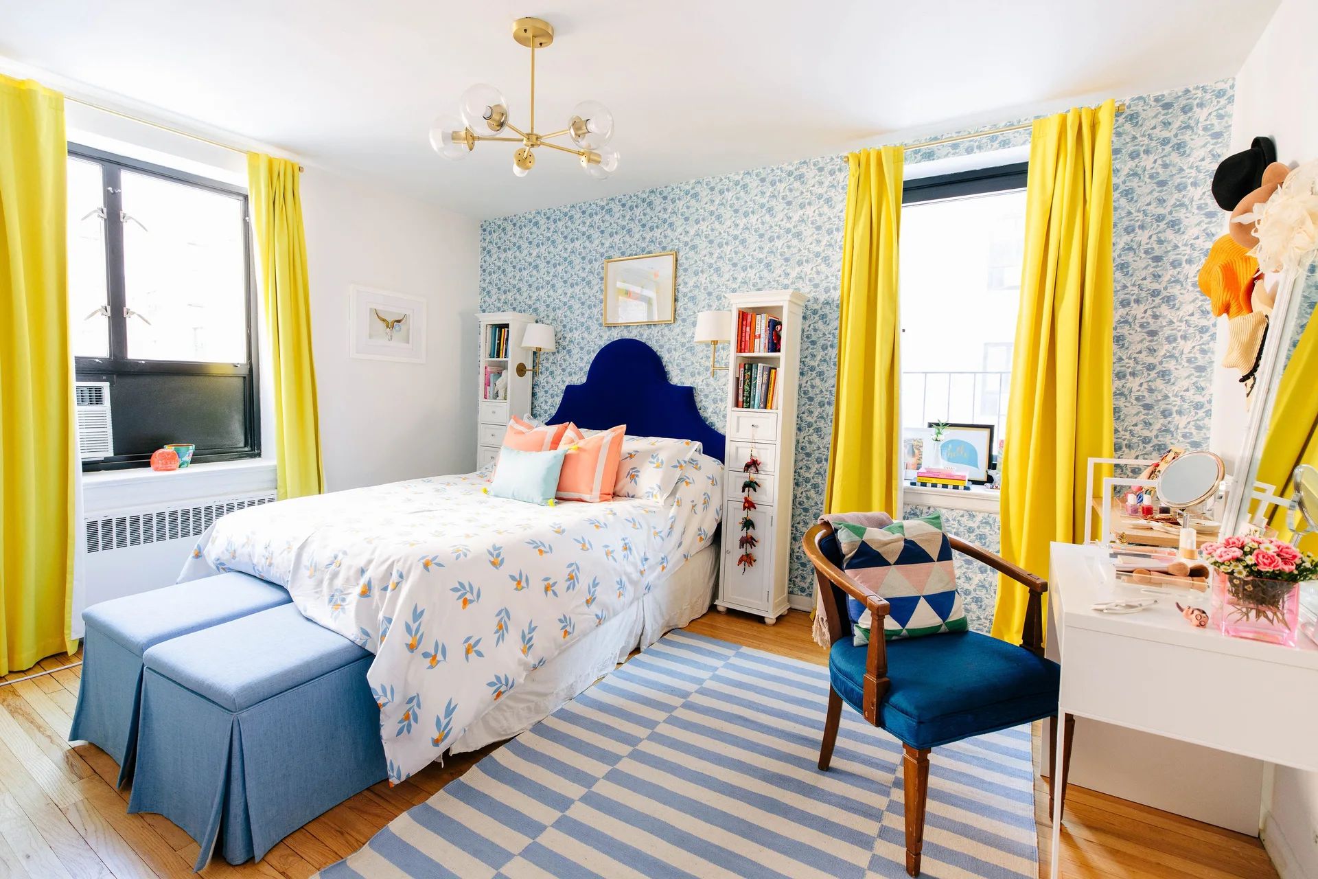 A bedroom features a blue arched headboard against floral wallpaper, flanked by bright yellow curtains, a striped rug, and a white vanity.