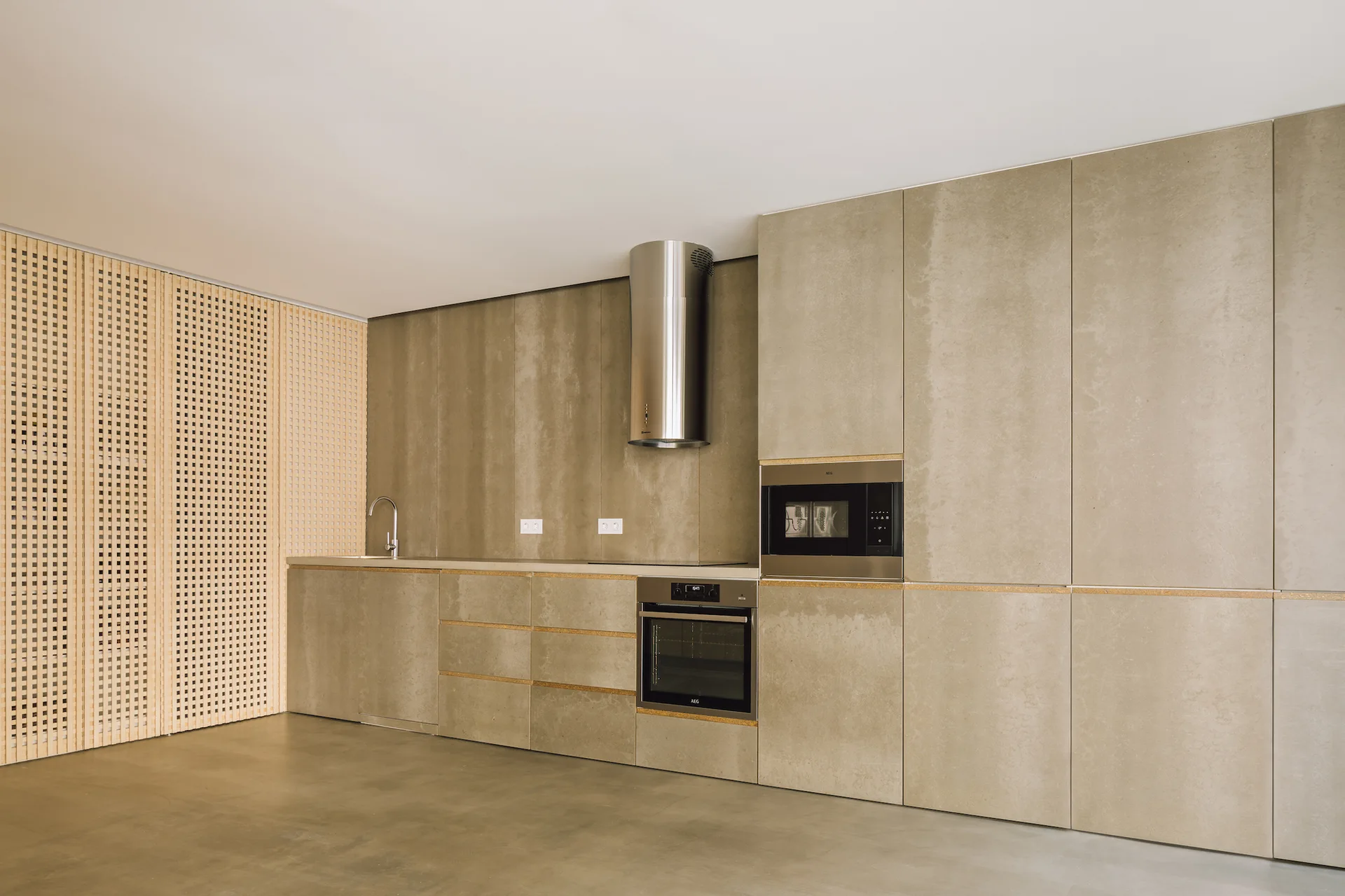 A minimalist kitchen features seamless taupe cabinetry, a cylindrical stainless steel range hood, and a light-wood lattice wall panel.